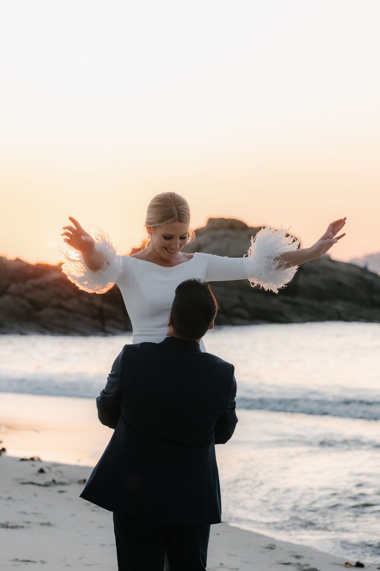 A couple shares a joyful dance on the beach at sunset with ocean waves in the background.
