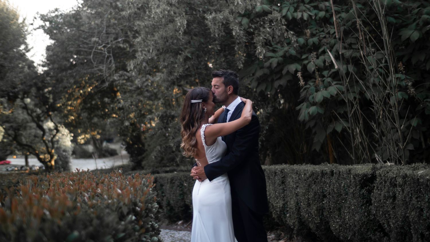 A couple embraces in an intimate moment at sunset against a backdrop of lush greenery and stone walls.
