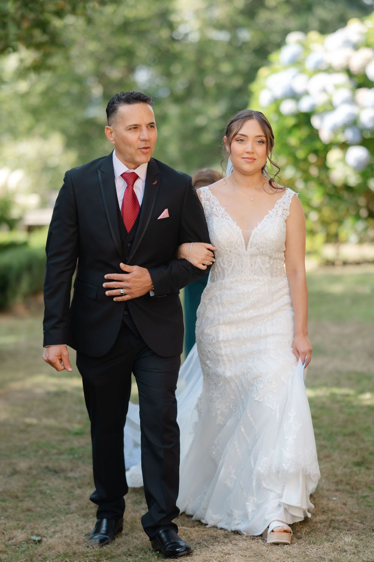 A couple walks arm in arm outdoors, with the bride wearing a white lace gown and the groom in a black suit with red tie.