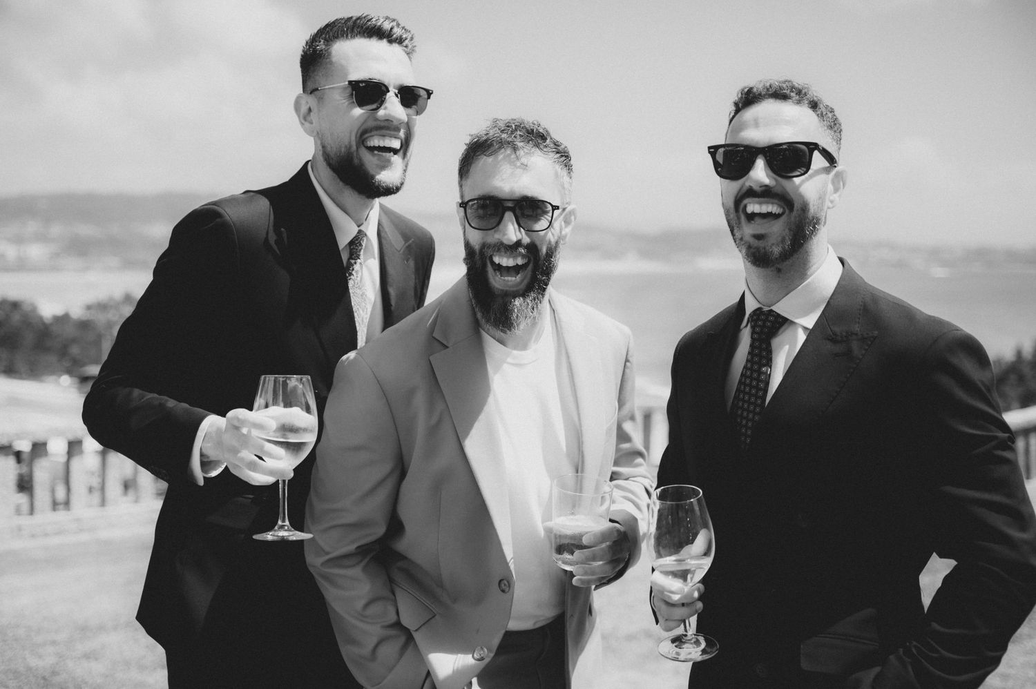 A group of groomsmen in black suits laugh and share drinks during an outdoor wedding celebration.