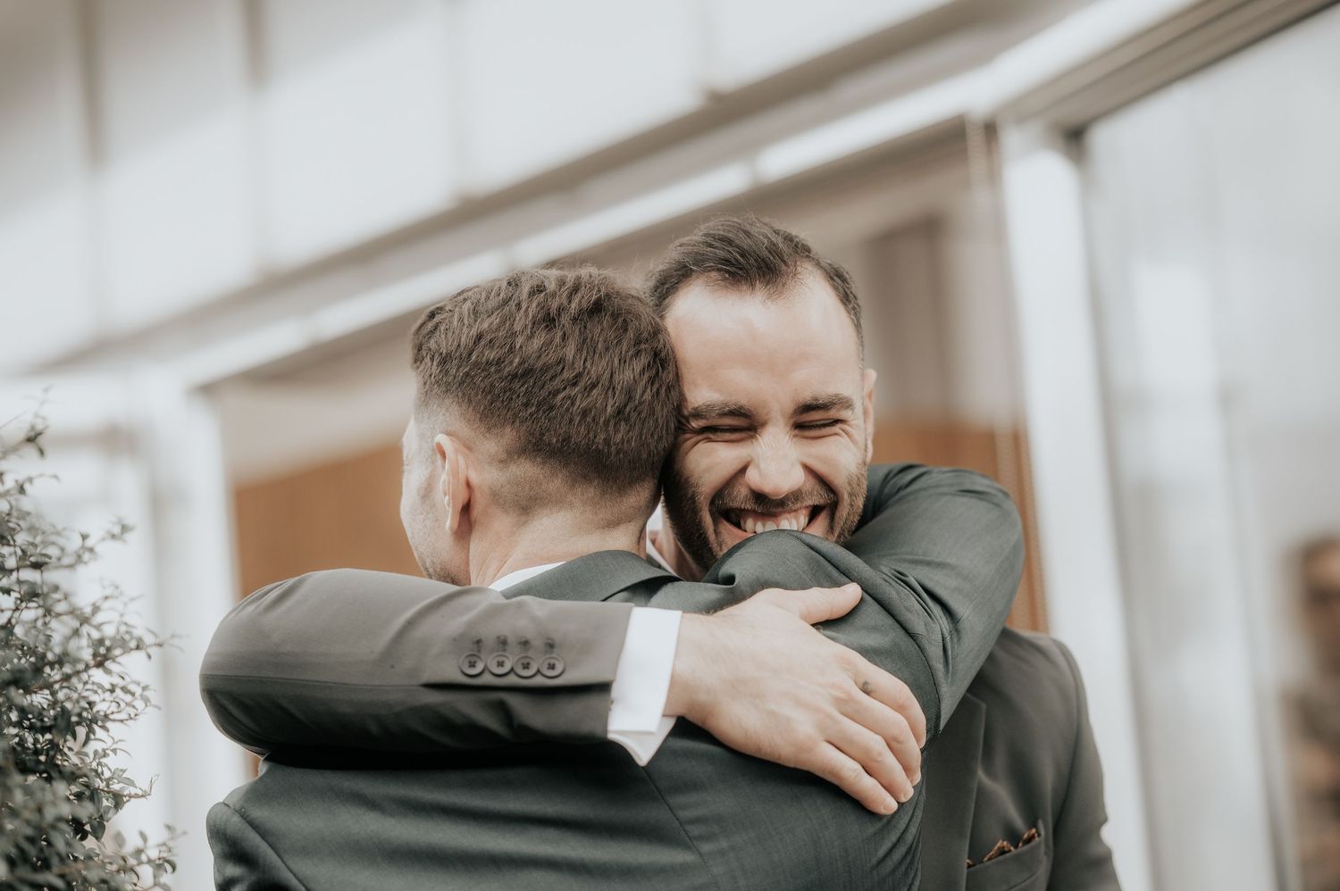 Two people share a joyful embrace outdoors on a bright day wearing formal suits.