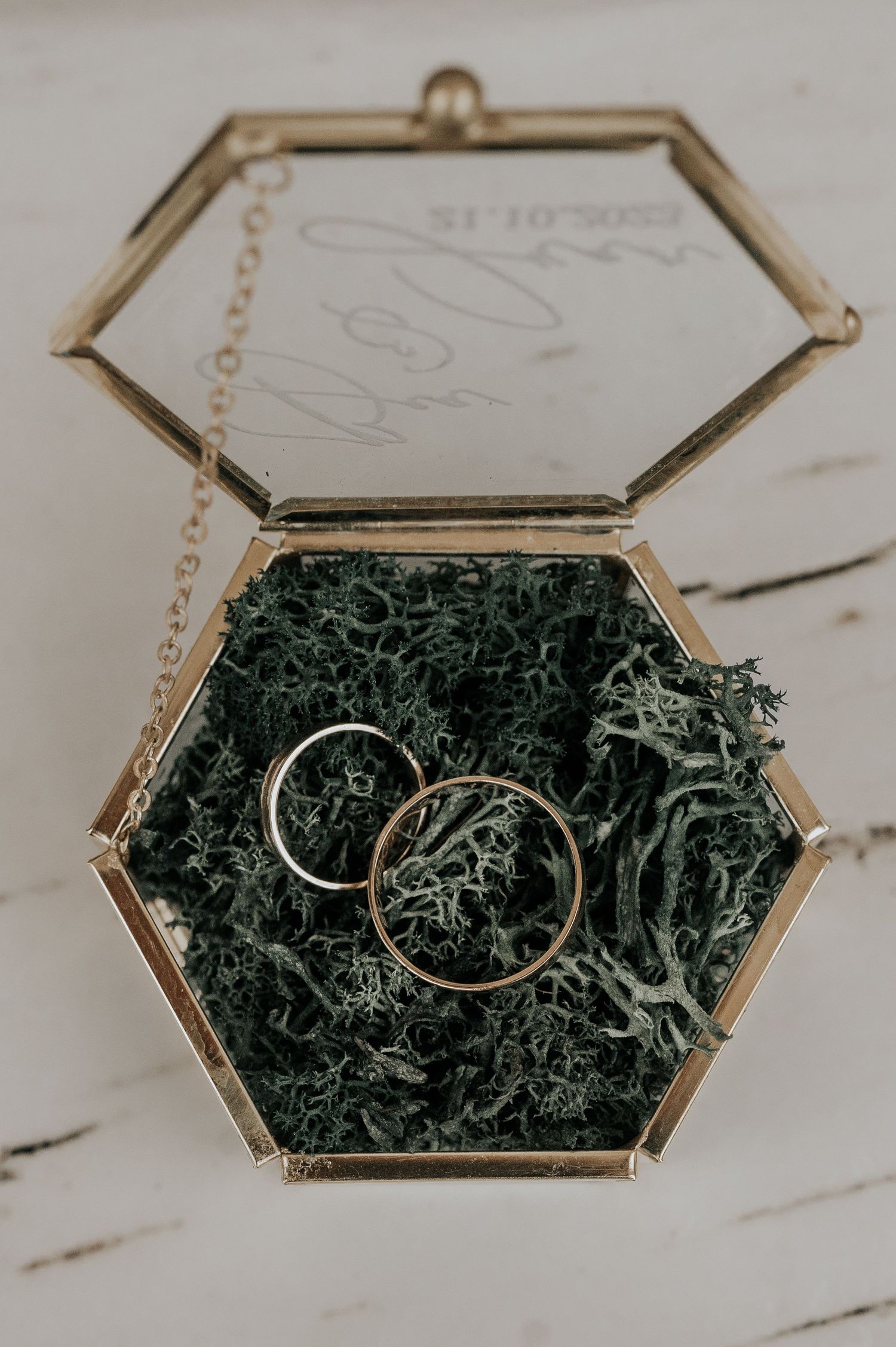 Wedding rings nestled in moss inside a hexagonal glass display box on a marble surface.