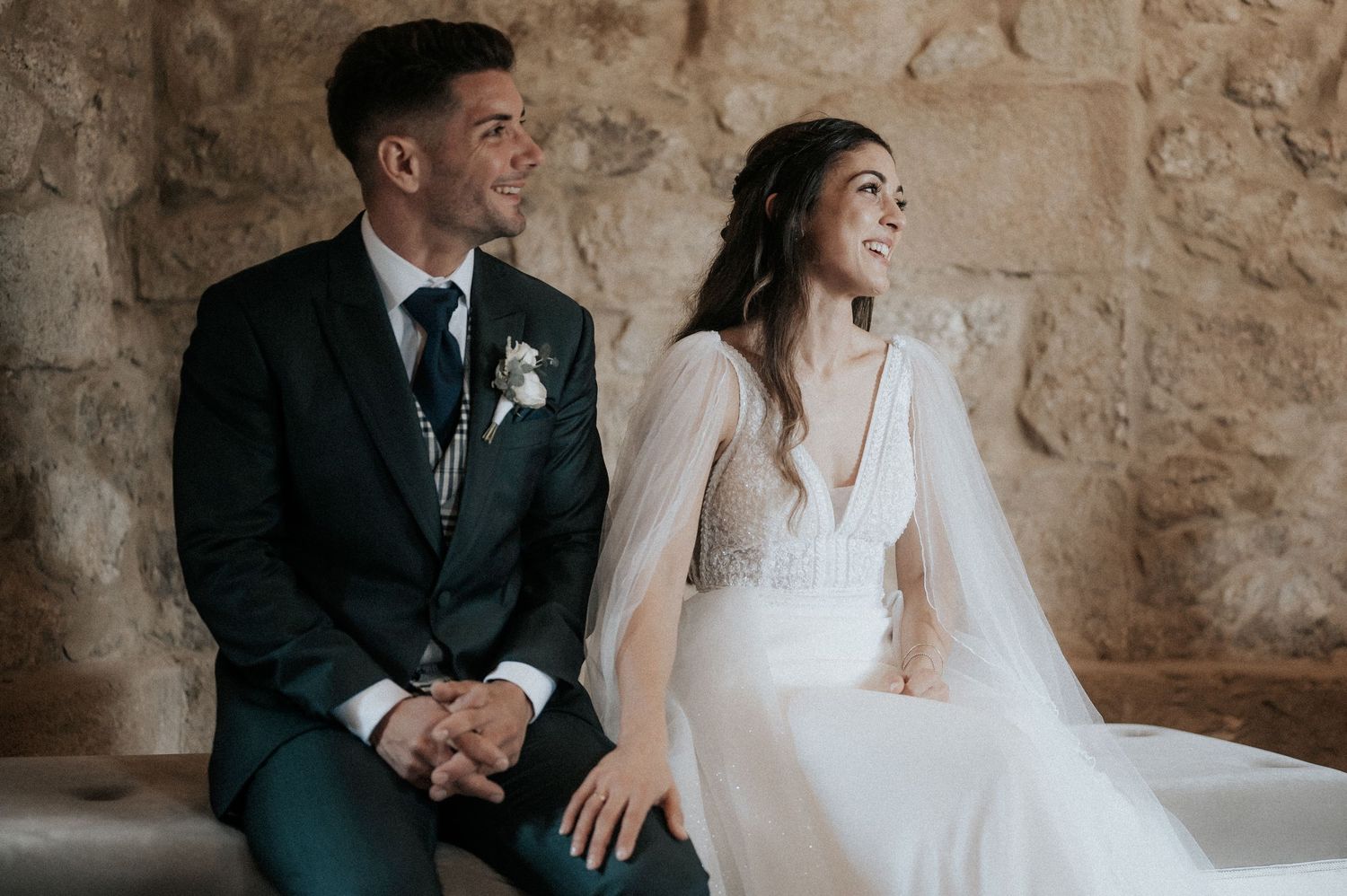 Newlywed couple sitting together against a rustic stone wall backdrop during their wedding photoshoot.