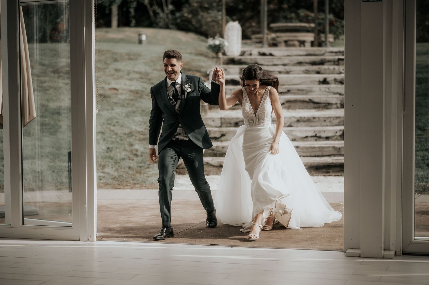 A couple runs playfully through a modern glass-walled building corridor during their wedding celebration.