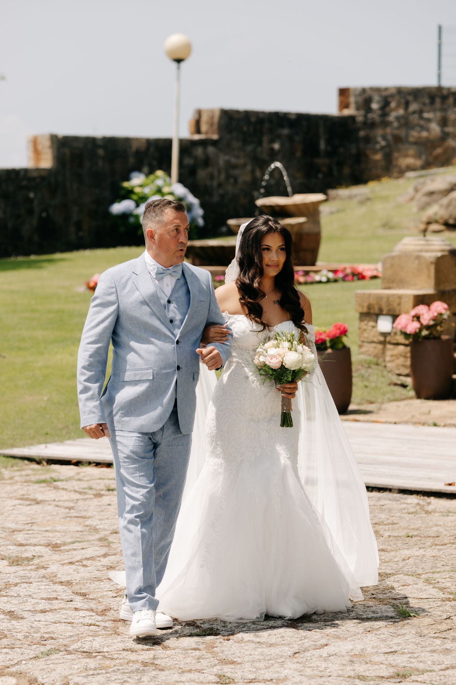 A couple walks together at a historic stone fortress on their wedding day, with pink flowers decorating the grounds.