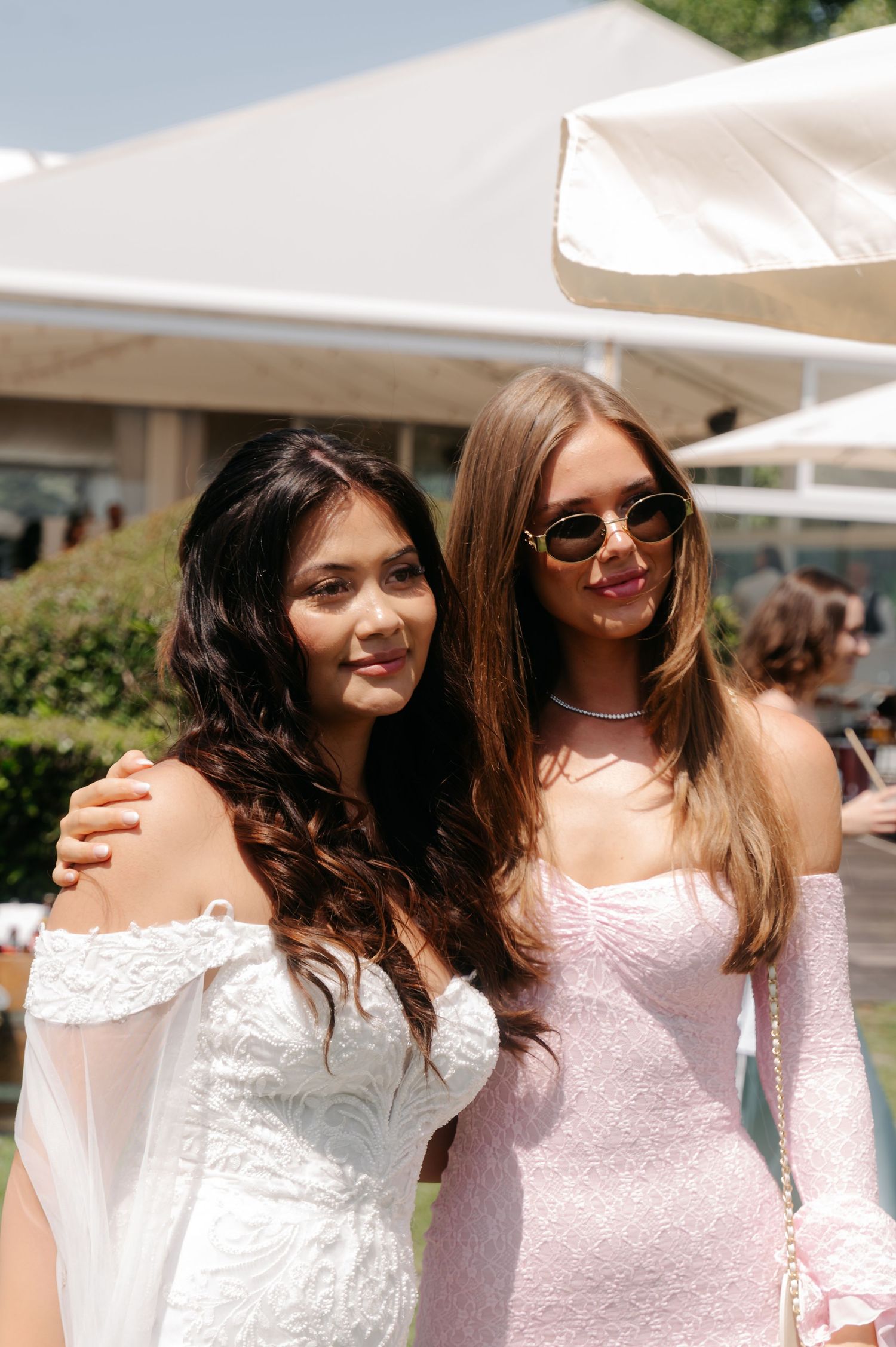 Two women in light colored dresses laugh and smile together at an outdoor event under white umbrellas.