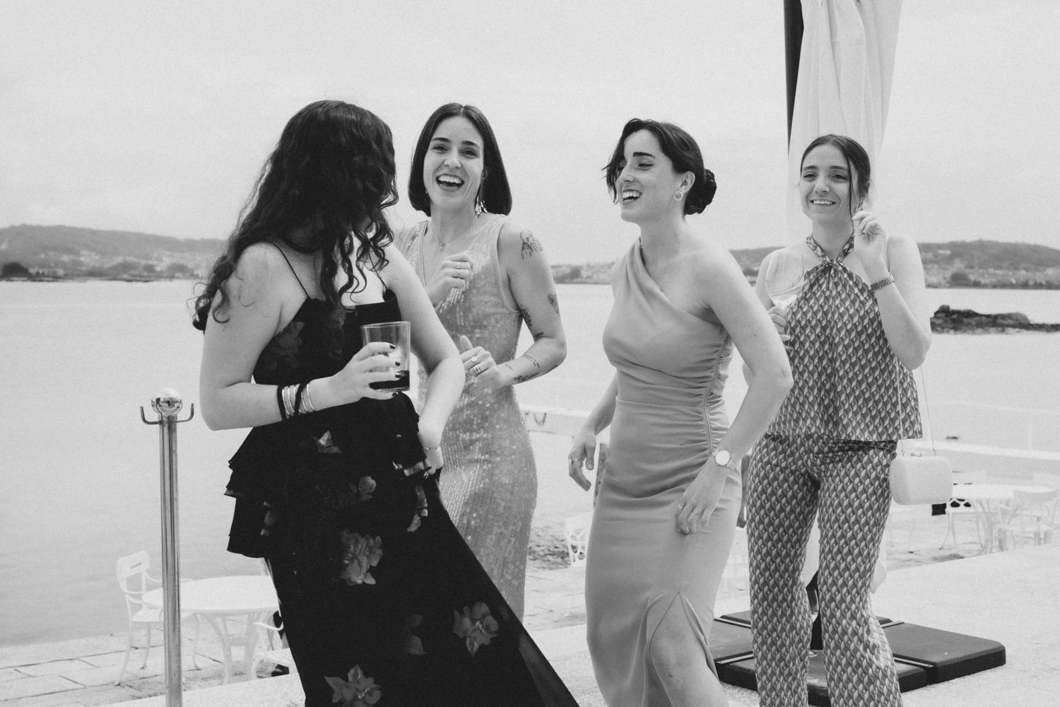 A group of wedding guests laughing and socializing on a sunny outdoor terrace by the ocean.