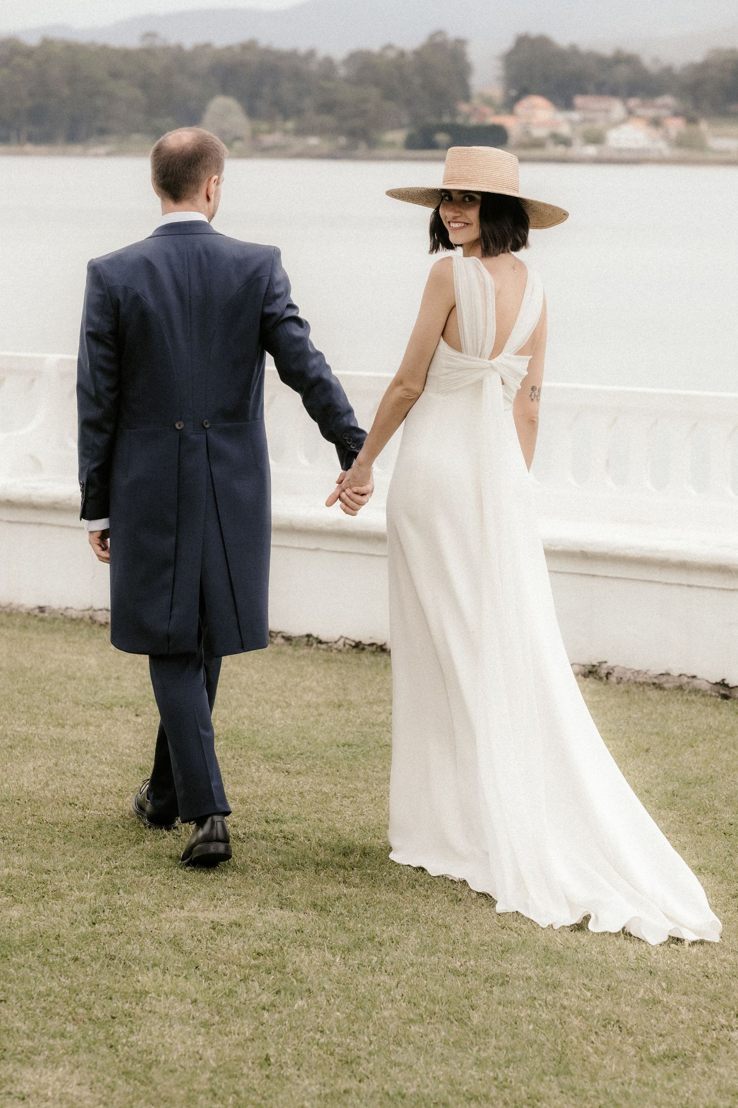 A couple in wedding attire walks along a waterfront path with scenic views.