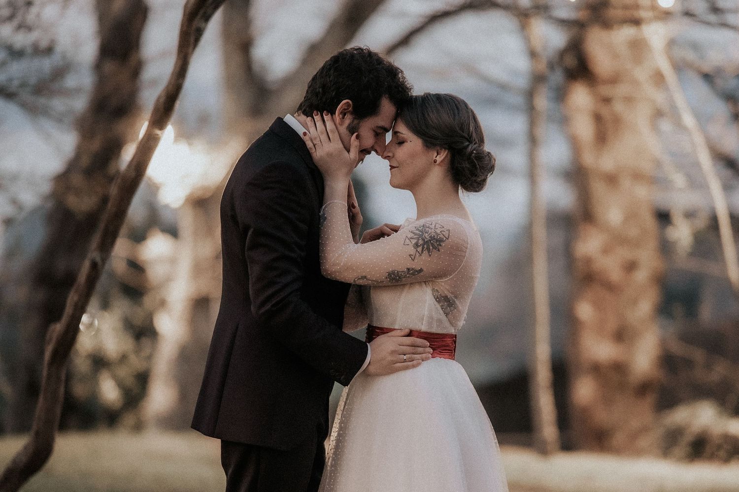 A romantic couple in wedding attire share an intimate moment together in a wooded outdoor setting at sunset.