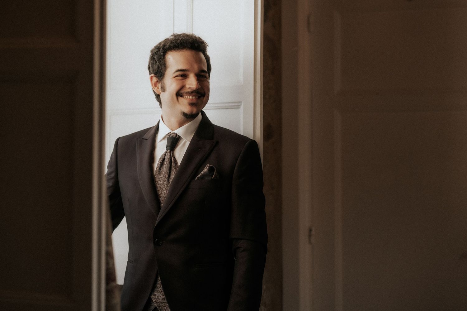 Groom in classic black suit laughs joyfully during wedding preparations in warm lighting.