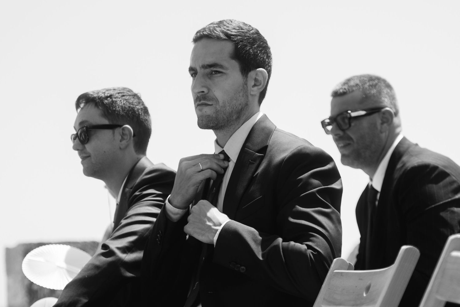 Three men in dark suits and sunglasses sit together in a formal setting, captured in black and white photography.