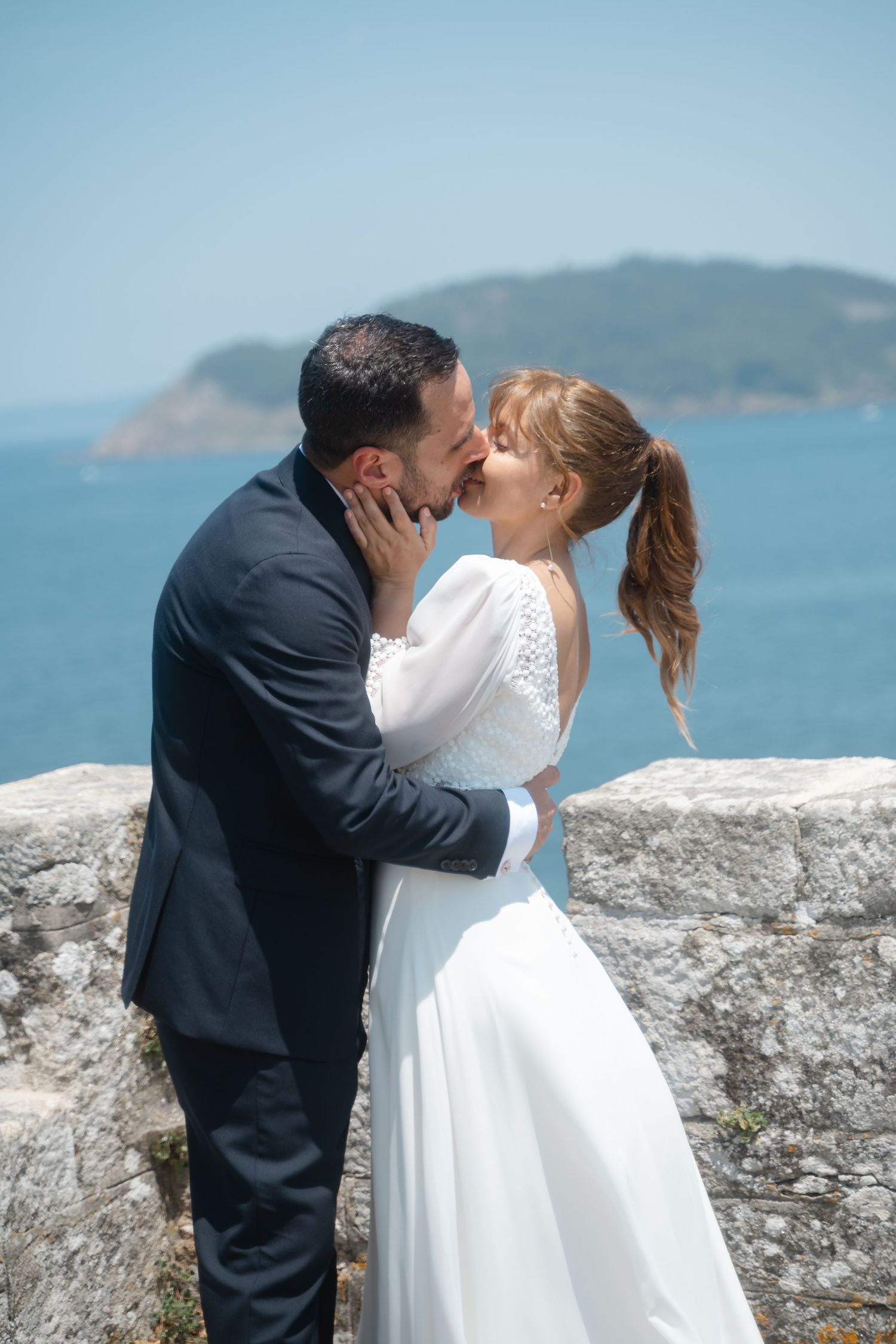 A romantic wedding couple shares a kiss on a coastal overlook with mountains and ocean in the background.
