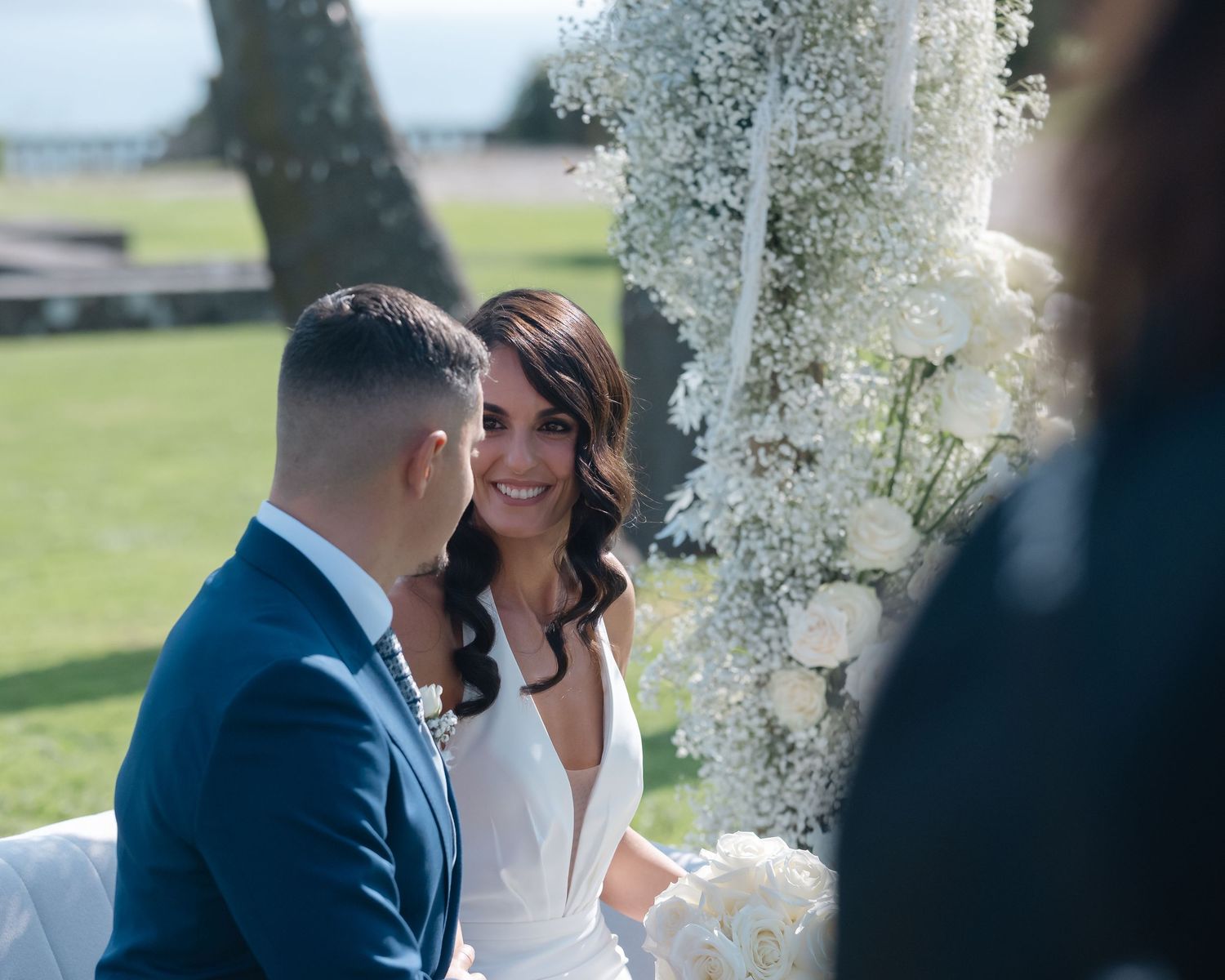 Newlyweds share an intimate moment under white floral decorations at an outdoor wedding ceremony.