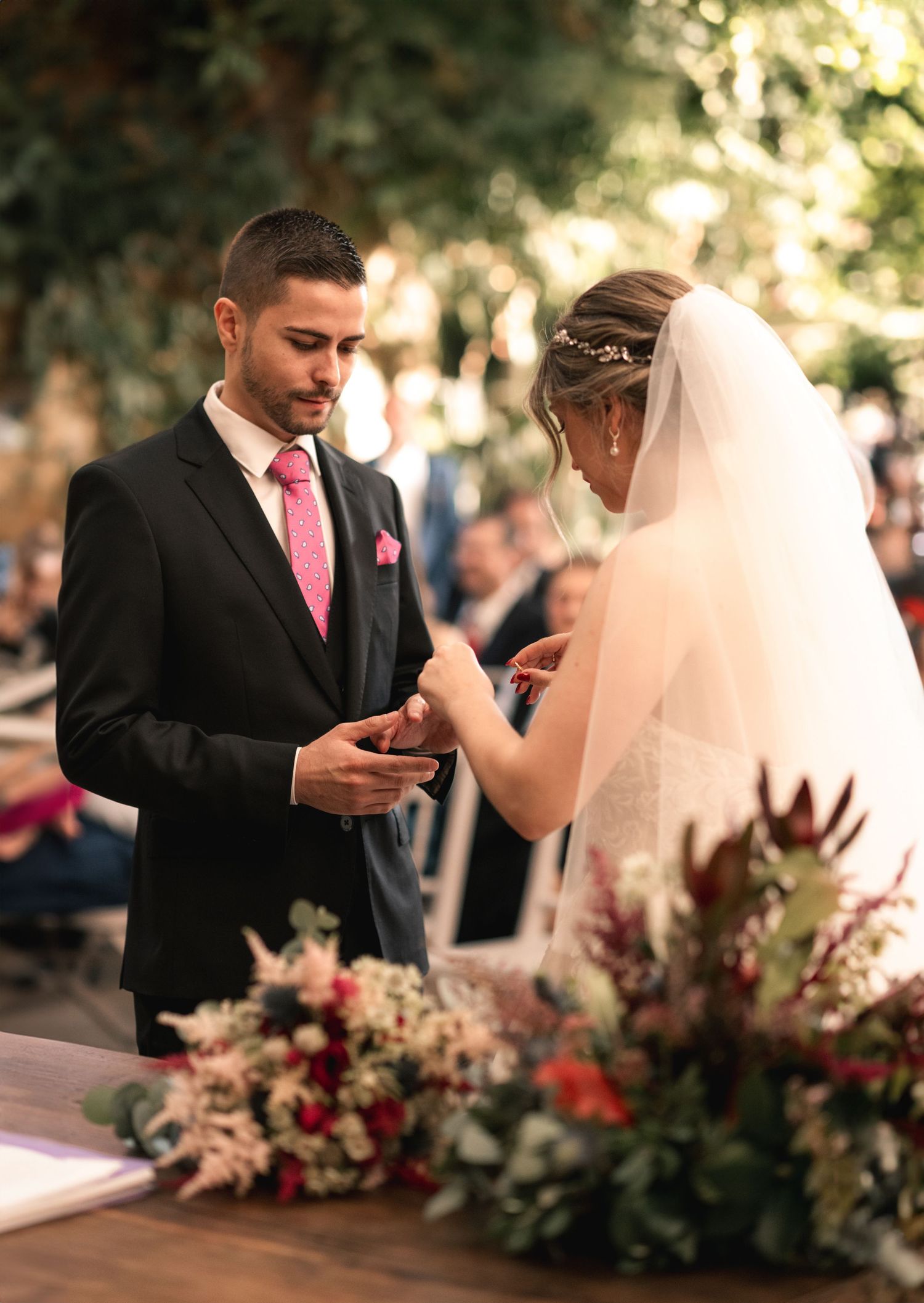 A wedding ceremony moment showing the exchange of rings with pink floral arrangements decorating the outdoor setting.
