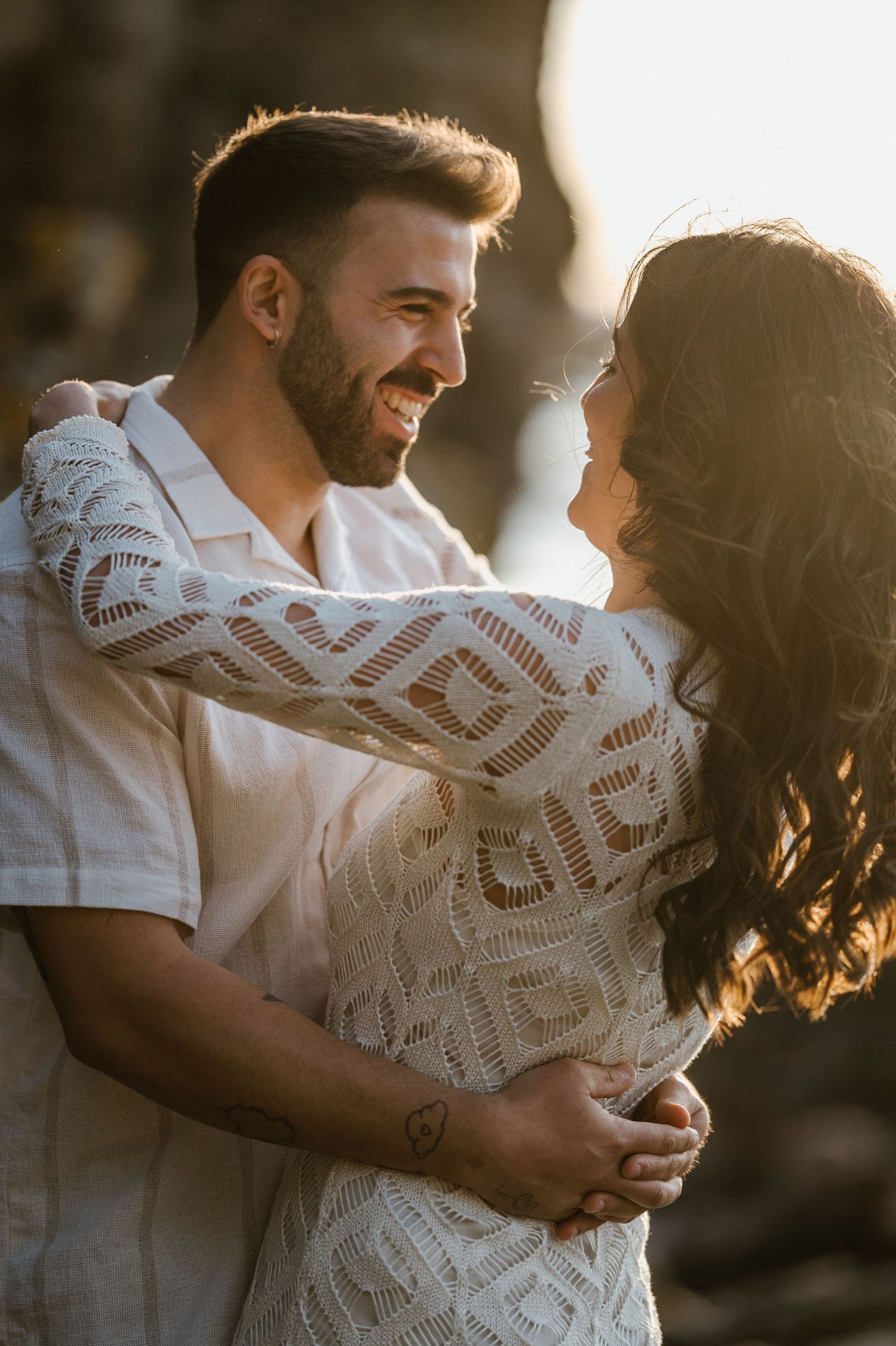 Romantic couple embraces in golden sunlight wearing white lace and casual attire during sunset photoshoot.