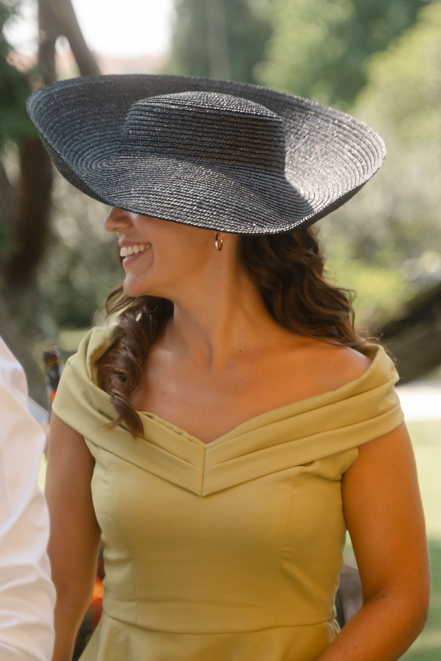 Wedding guest wearing a stylish yellow dress and wide-brimmed hat smiles during outdoor celebration.