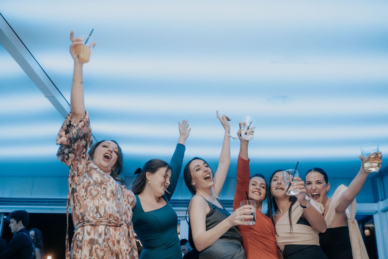 Group of friends enjoying drinks and dancing outdoors against a blue sky background.
