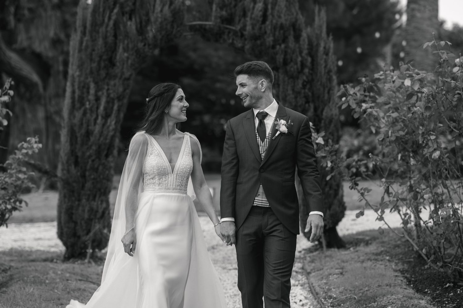 Wedding couple walks through scenic garden path with tall cypress trees.