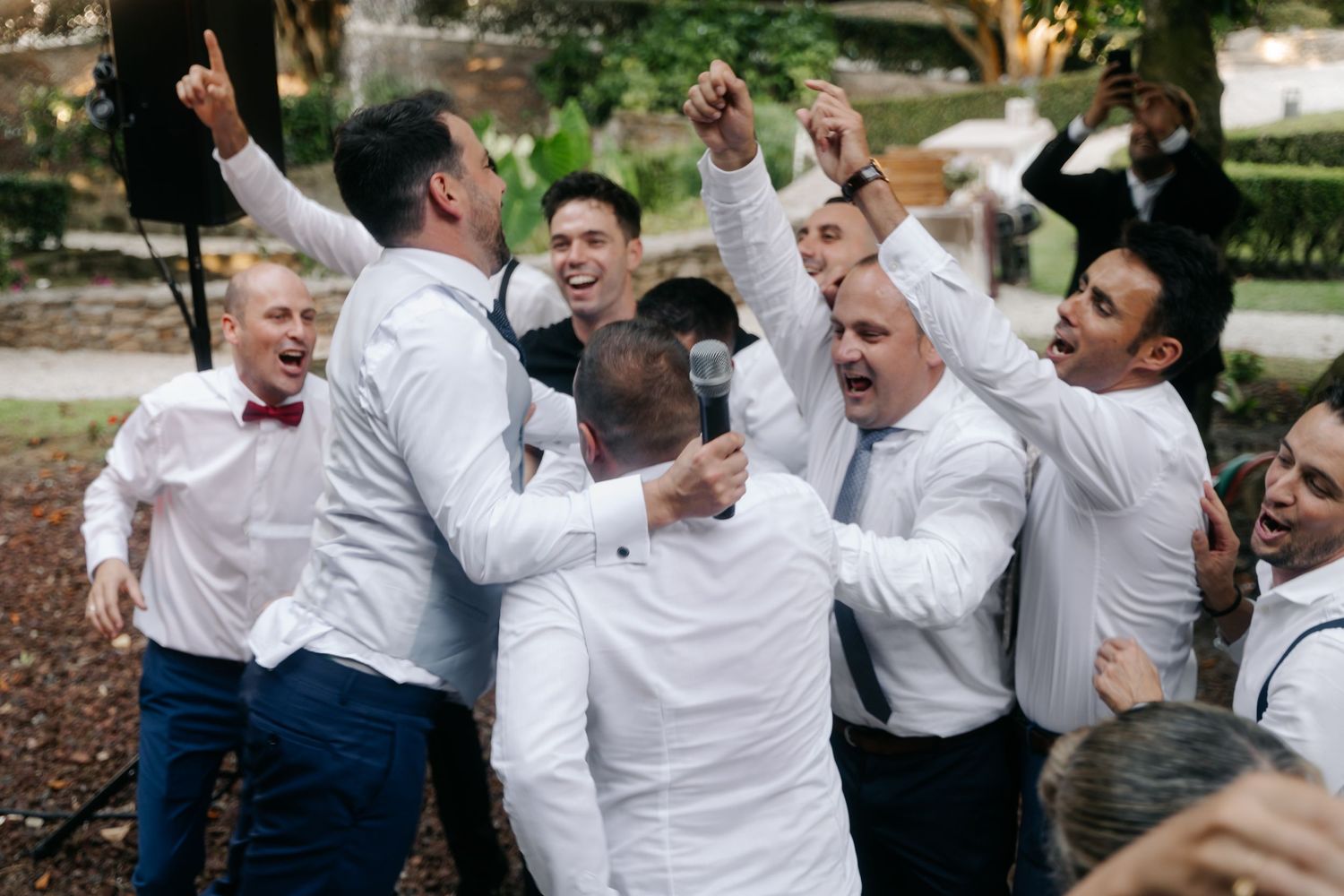 A group of groomsmen in white dress shirts celebrate joyfully together outdoors during a wedding event.