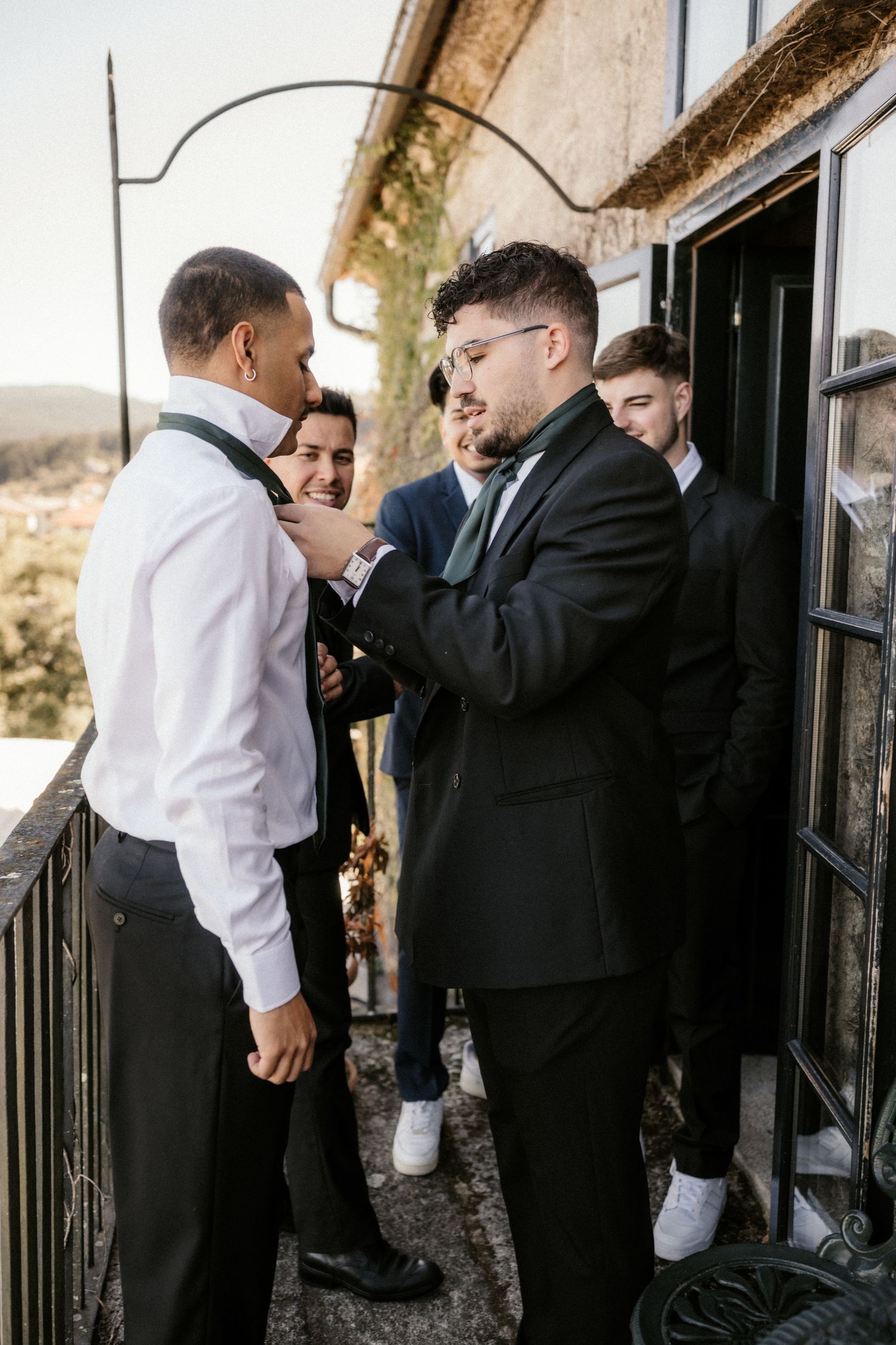 Groomsmen share a lighthearted moment while getting ready on a balcony before a wedding ceremony.