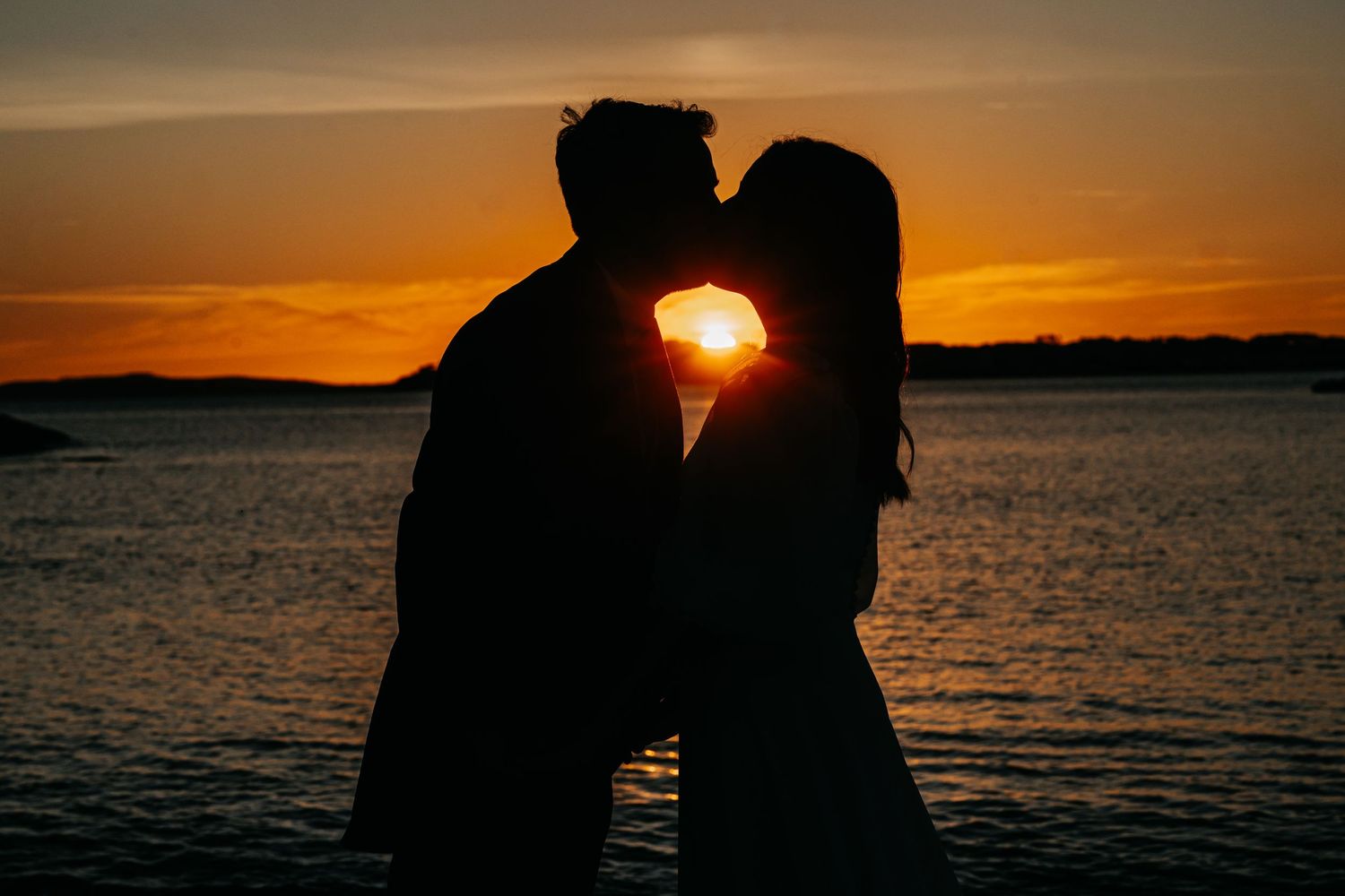 Silhouetted couple embracing against a vibrant orange sunset over calm ocean waters.