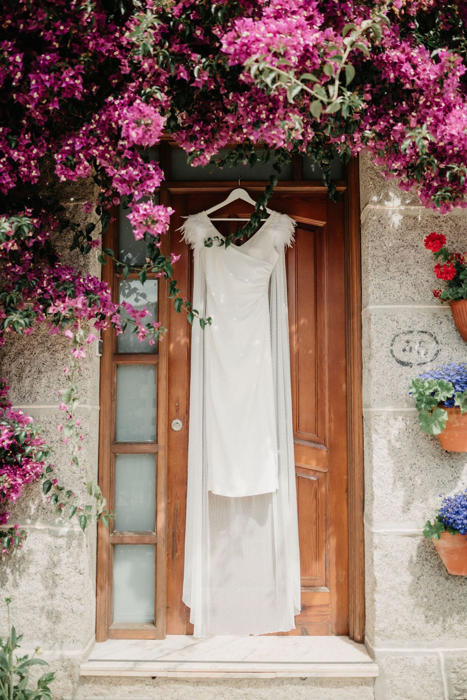 A white dress hangs on a rustic stone wall adorned with vibrant pink bougainvillea flowers and wooden windows.