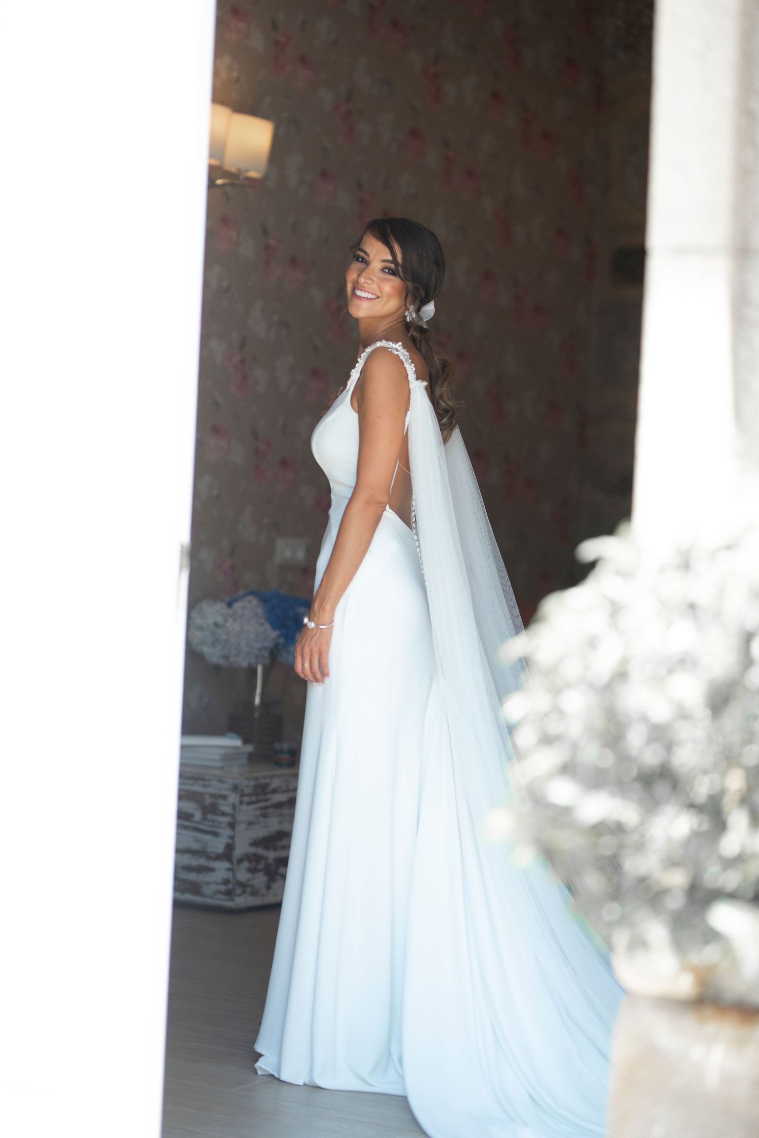 A bride in an elegant white wedding gown and veil poses between white columns in a romantic setting.