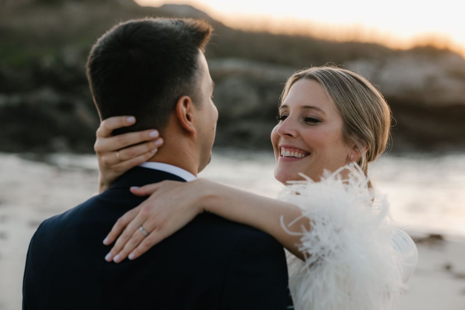 Golden hour wedding portrait with couple embracing on rocky beach.