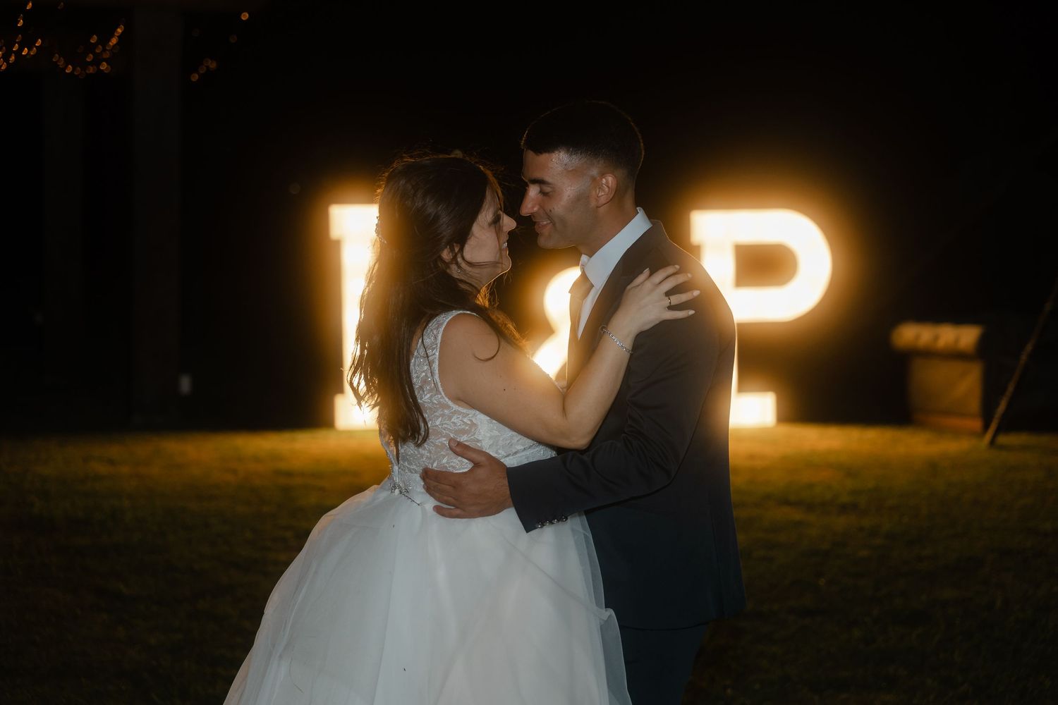 A romantic couple embraces and kisses at night with illuminated letters glowing in the background.