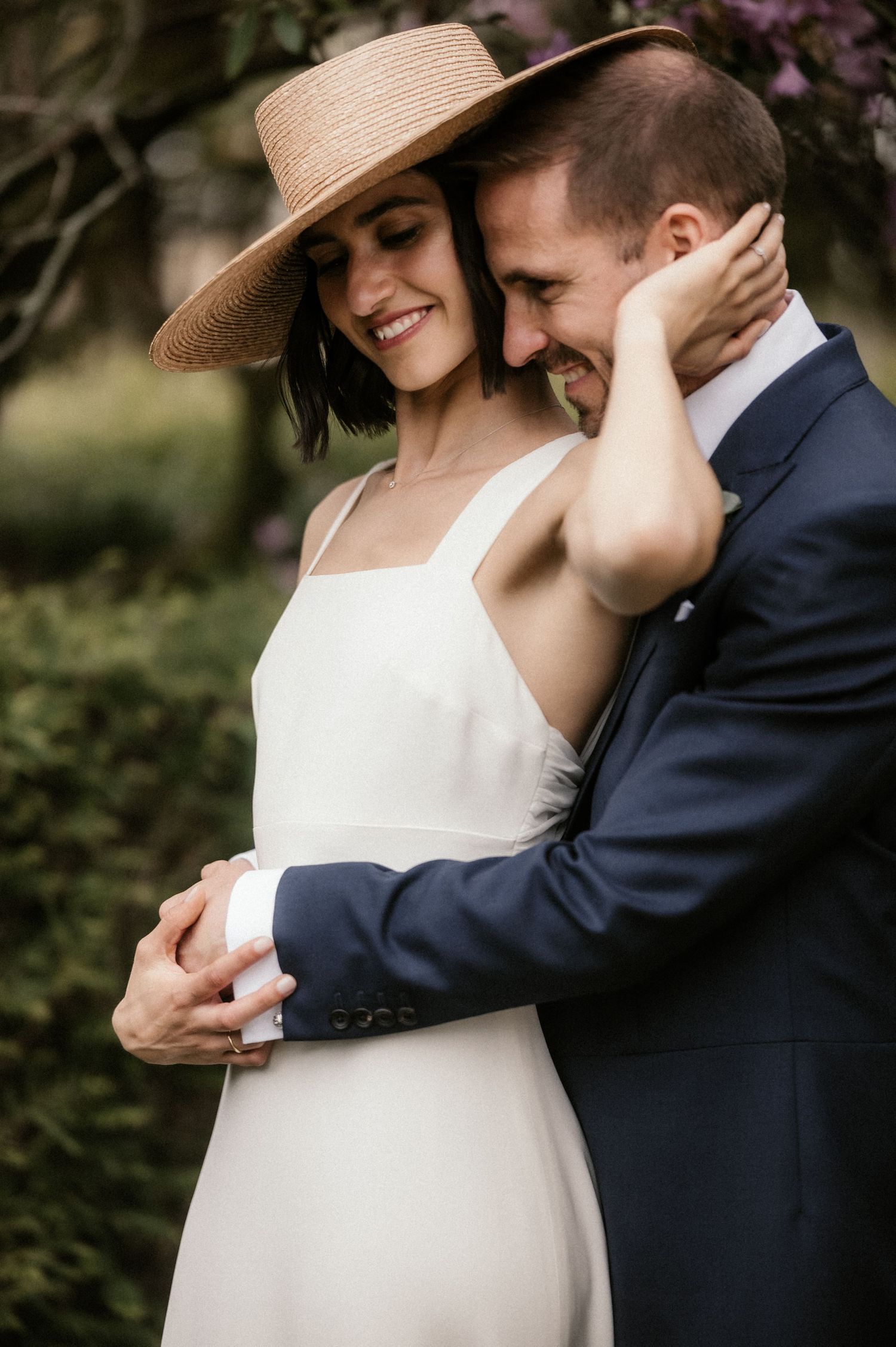 Intimate wedding couple embraces in a garden setting with the bride wearing a stylish wide-brimmed hat.