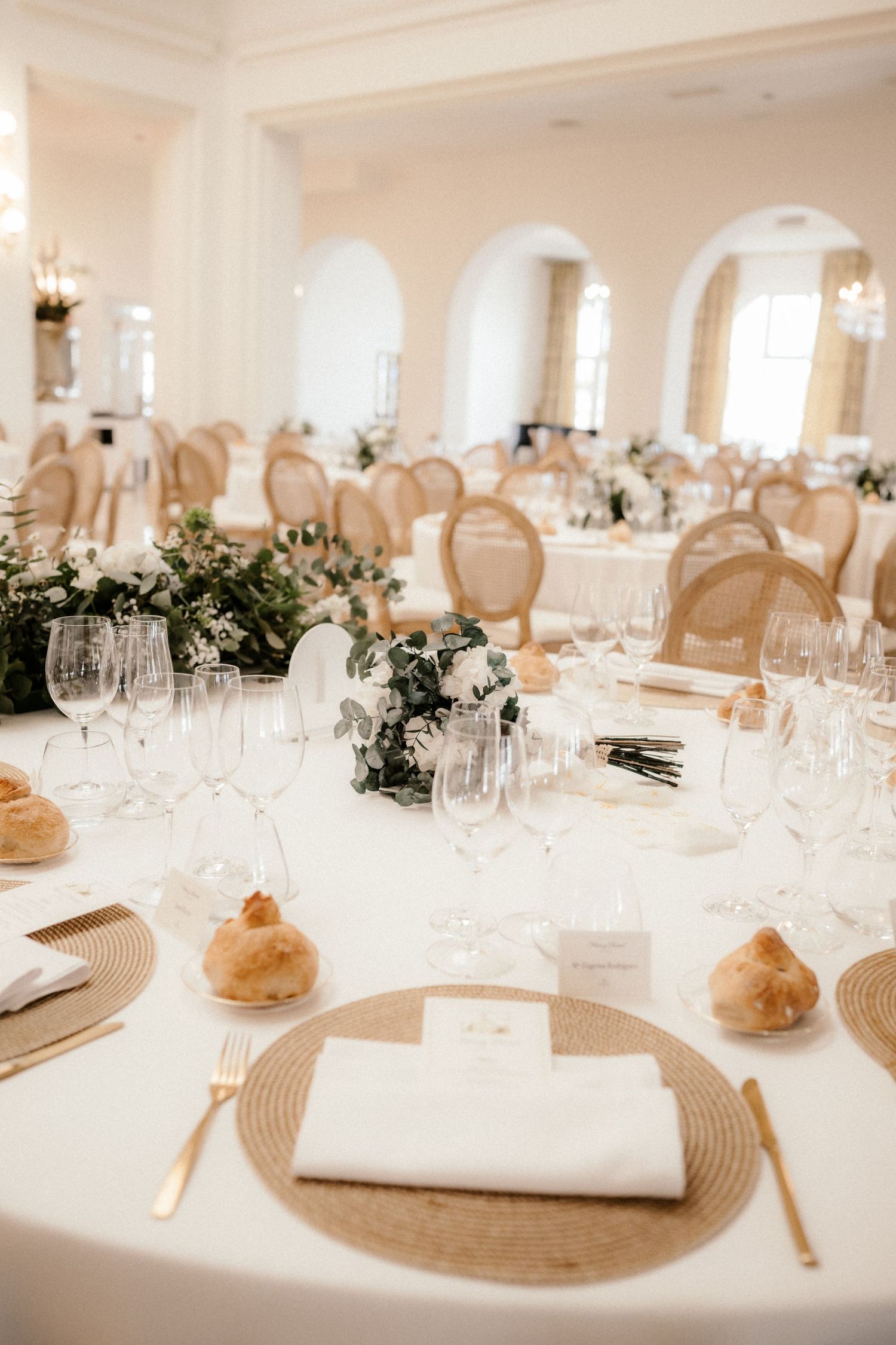 Elegant white wedding reception tables with crystal glassware, fresh greenery, and rustic bread rolls at each place setting.
