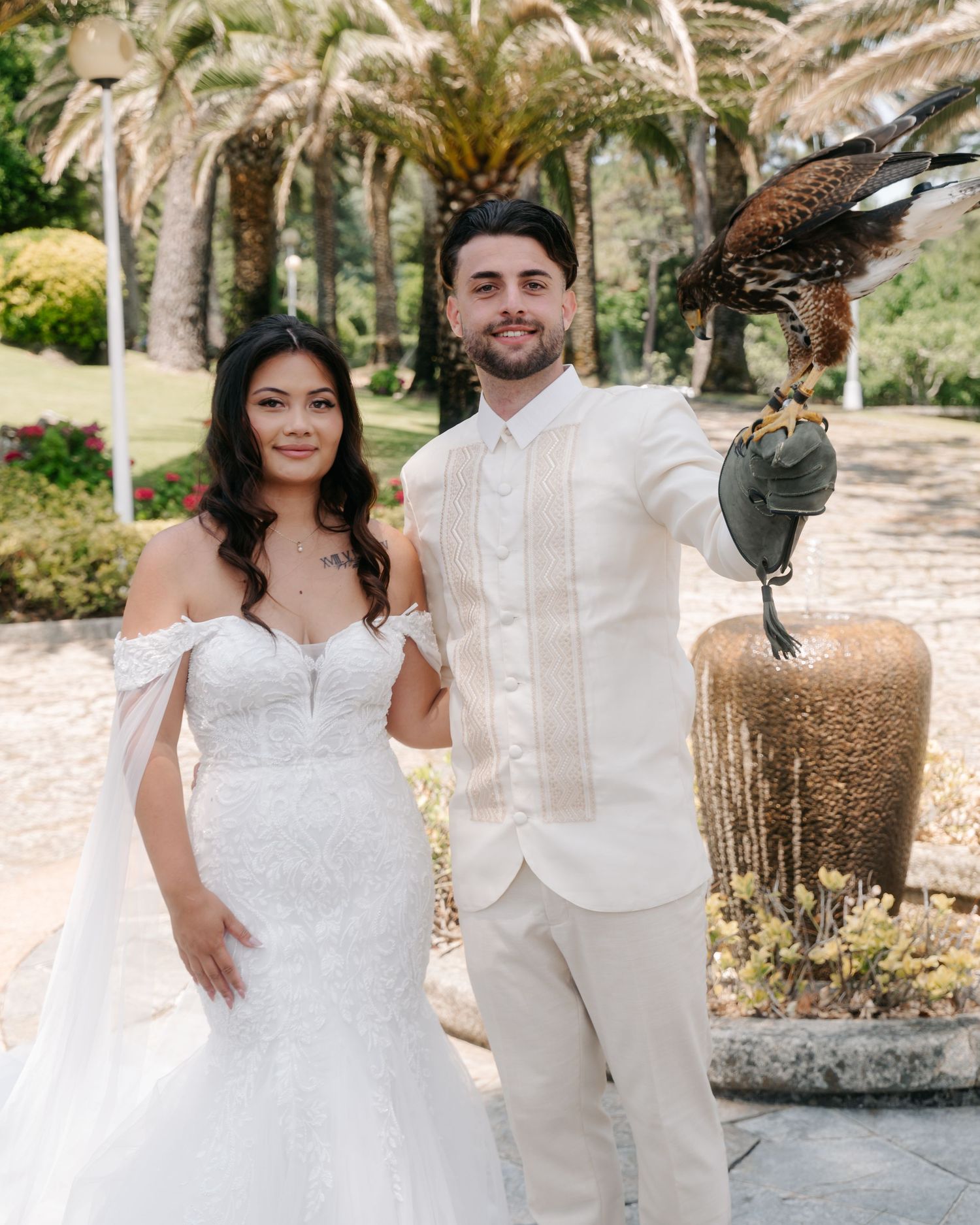 A couple poses outdoors at their wedding with palm trees in the background and a falcon perched on a stand.