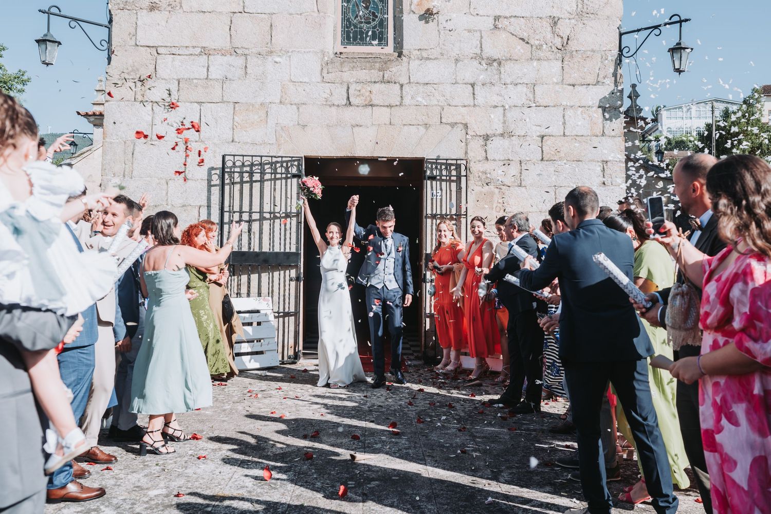 Wedding guests celebrate and dance together in front of a rustic stone building on a sunny day.