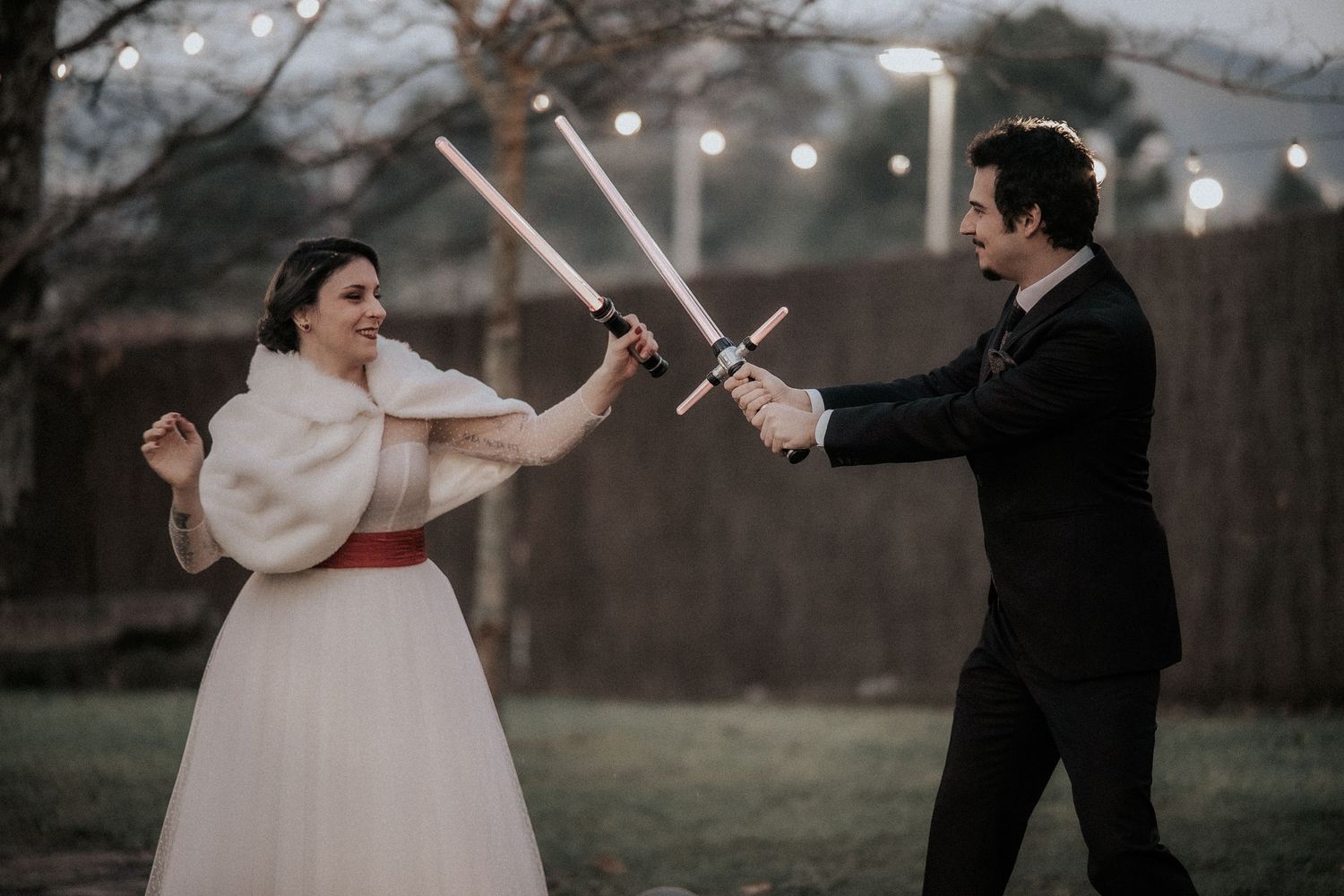 A couple in wedding attire playfully sword fights in a romantic outdoor setting at dusk with string lights in the background.
