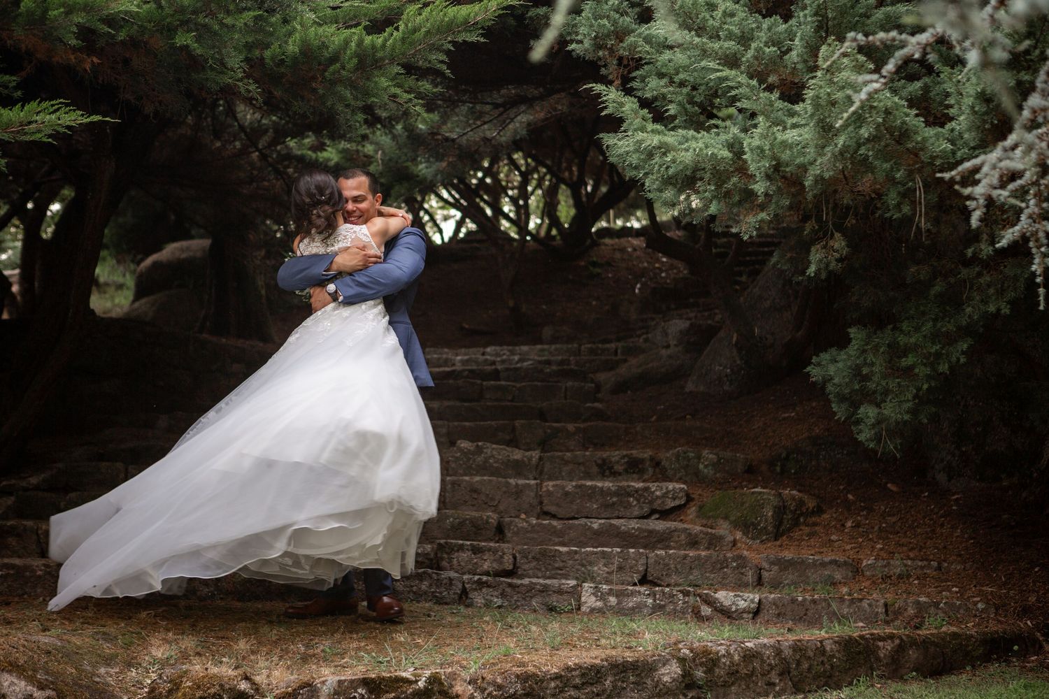 A bride in a flowing white wedding dress walks up rustic stone steps surrounded by lush greenery and trees.