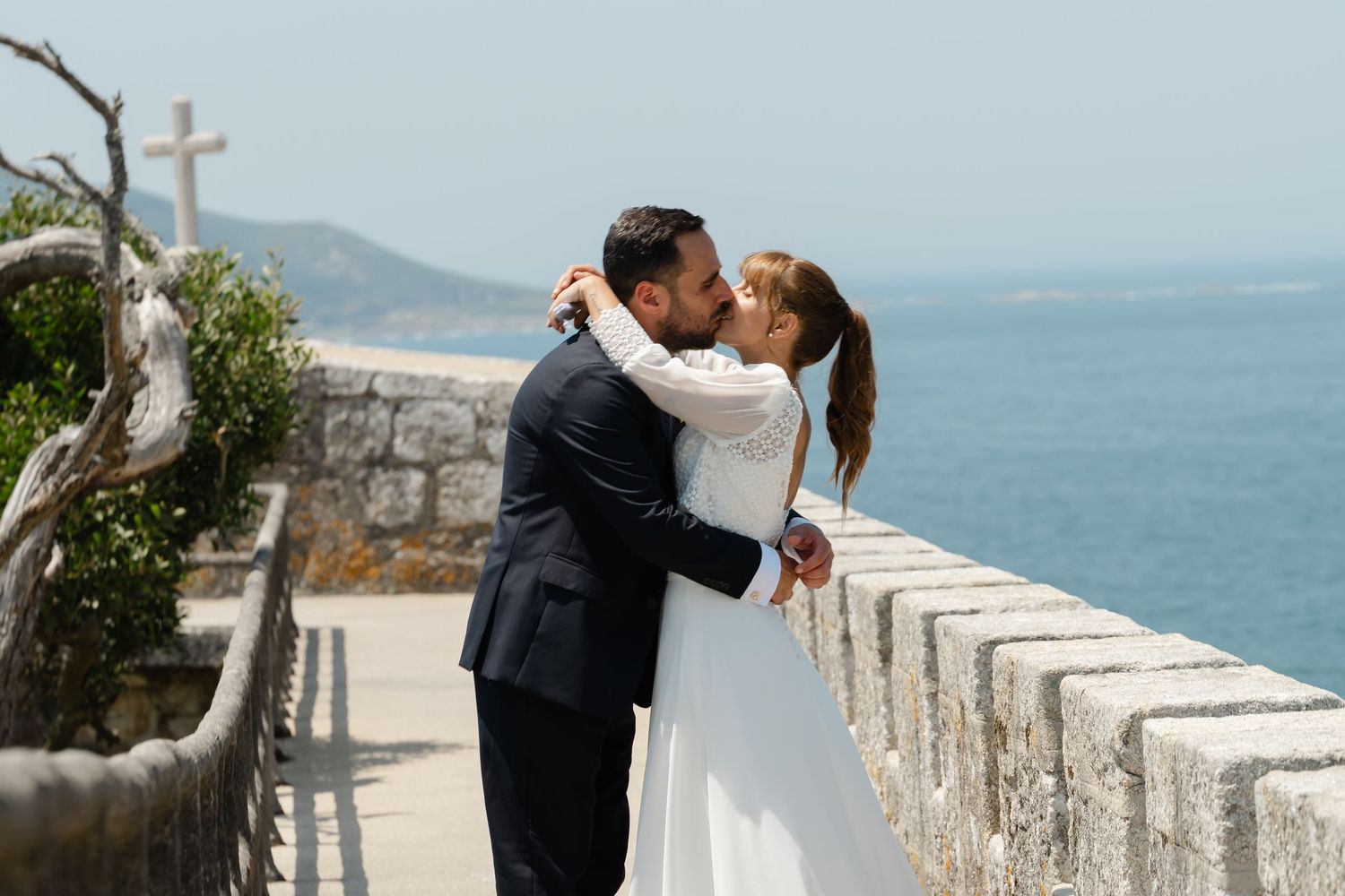 A couple shares a romantic kiss on a scenic coastal overlook with ocean views and stone walls.