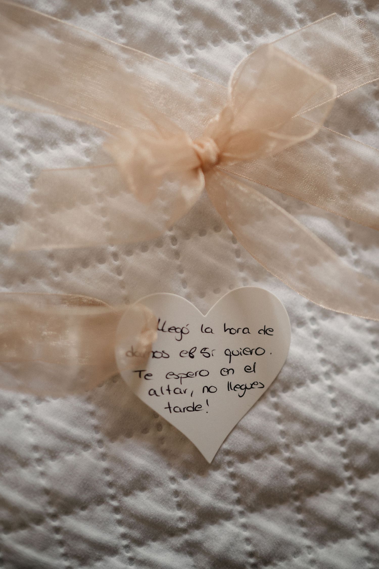 A delicate heart-shaped note with Spanish text is tied with a peach-colored tulle ribbon against a textured white surface.