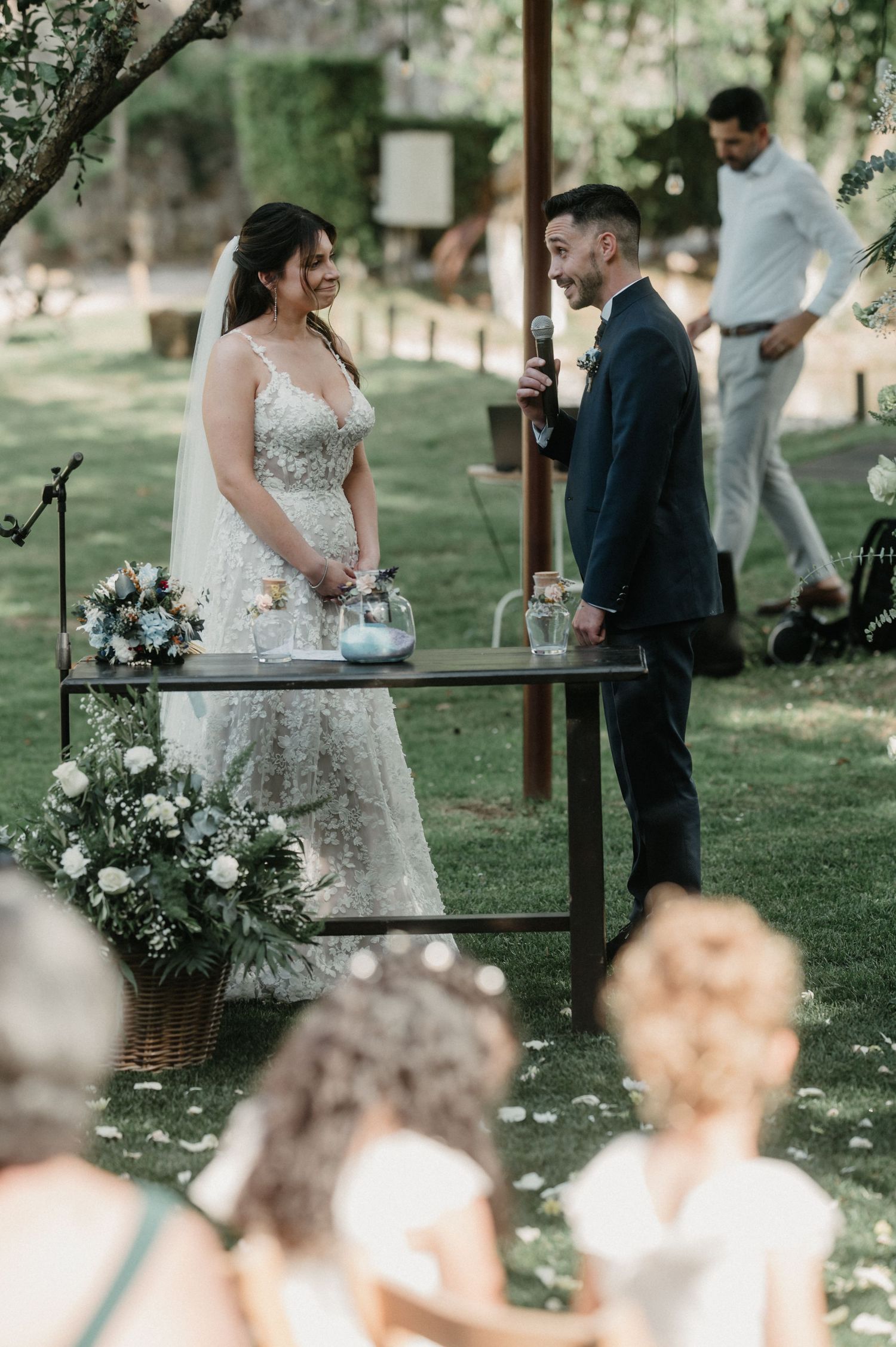 A couple stands at a rustic outdoor wedding ceremony table decorated with white floral arrangements in a garden setting.