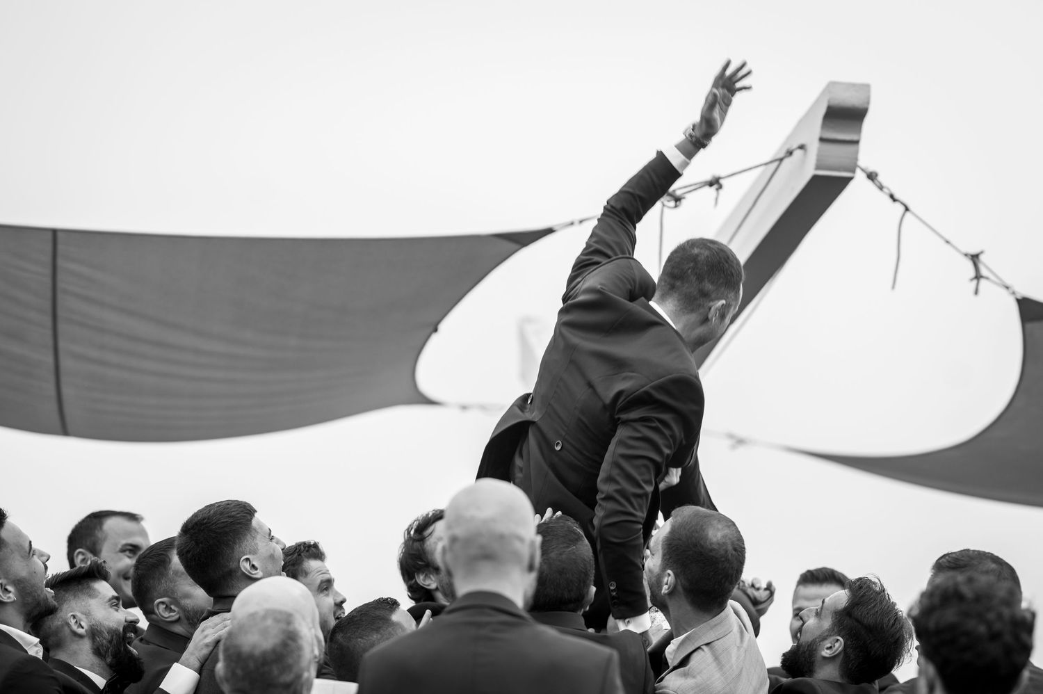 A crowd lifts someone up in celebration against a backdrop of a large flag in black and white.