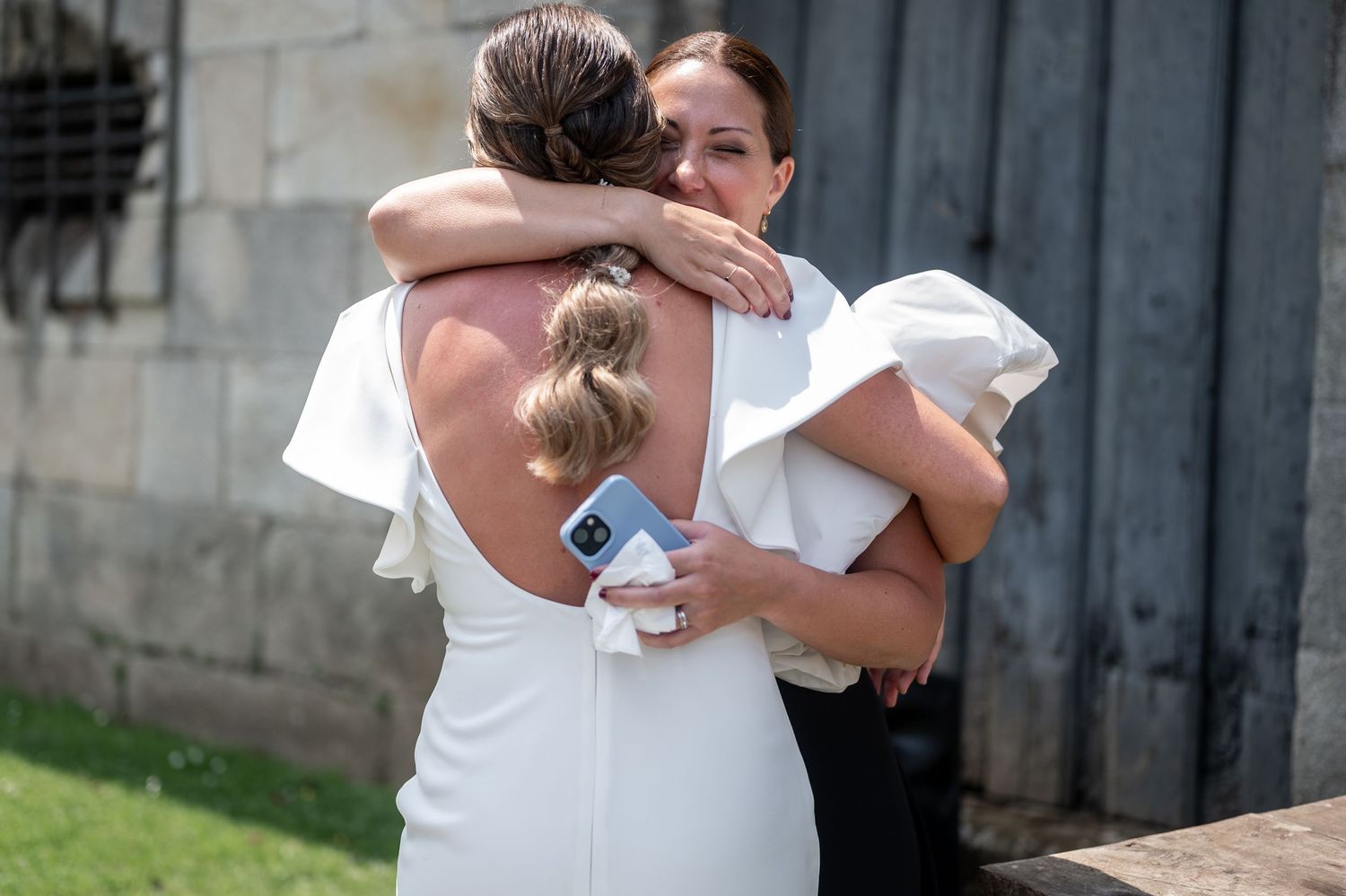 Two people in white shirts share an emotional embrace outside a stone building.