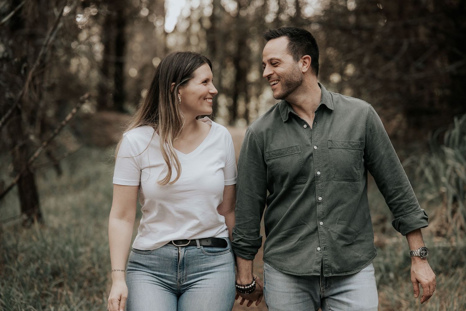 A couple walking together and smiling at each other in a wooded area during a casual outdoor photoshoot.