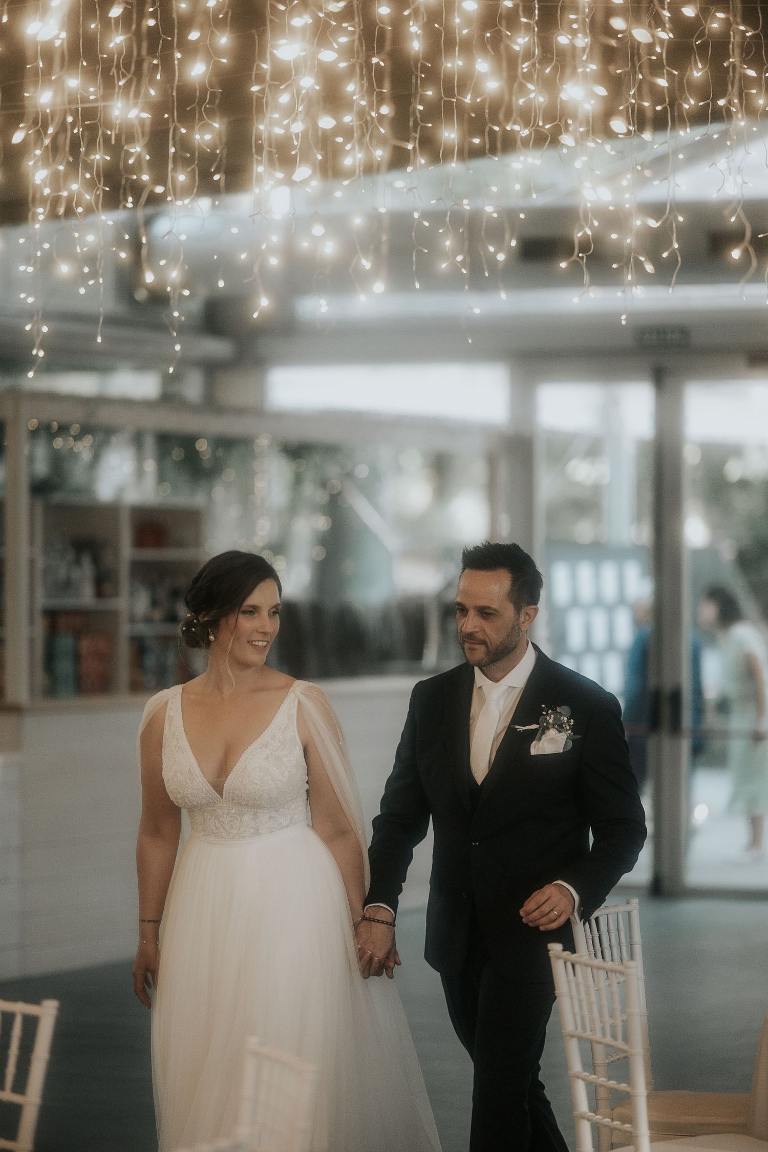A couple in wedding attire walks together under twinkling lights in an elegant indoor venue.