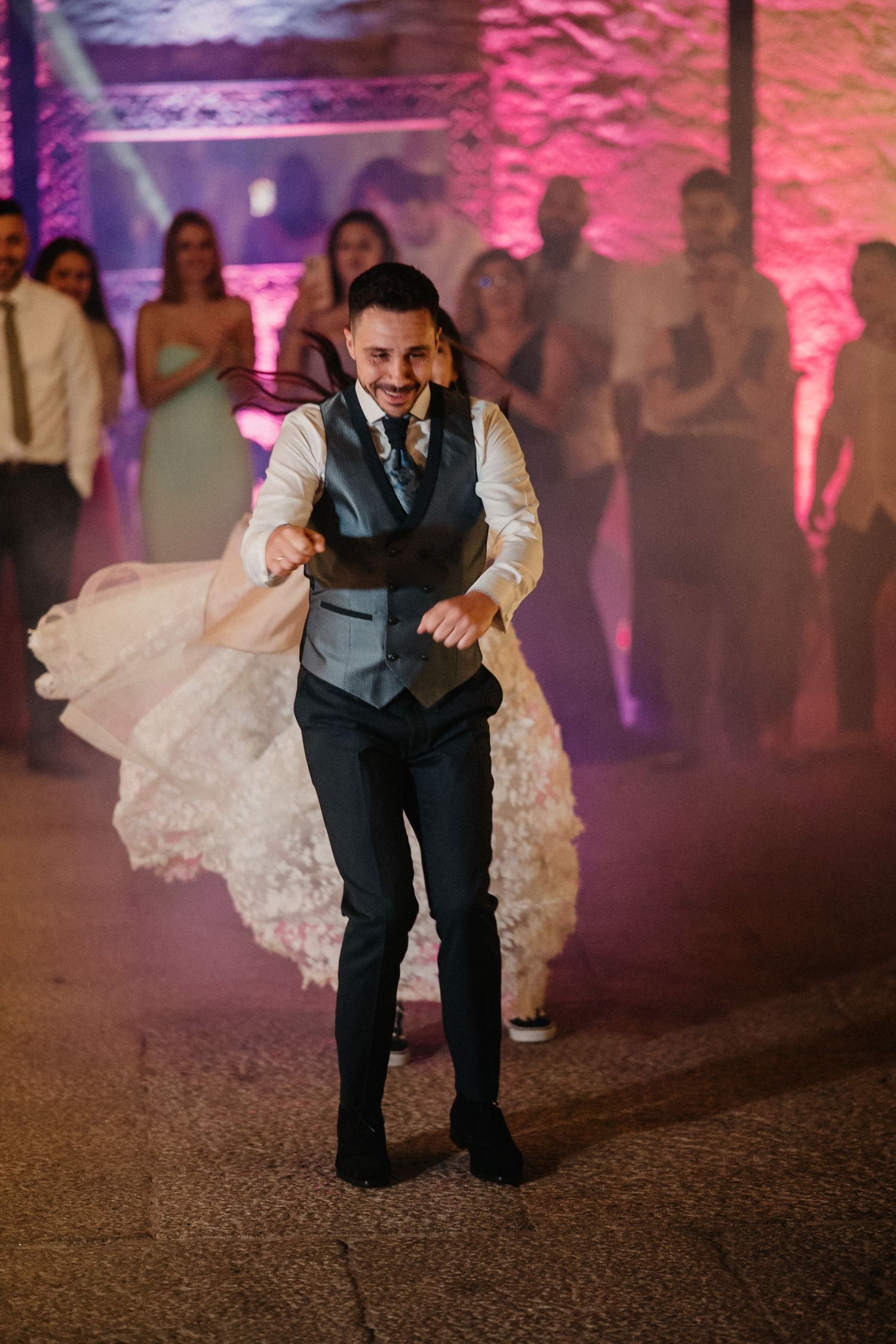Wedding party dancing under dramatic pink lighting at an indoor venue with stone walls.