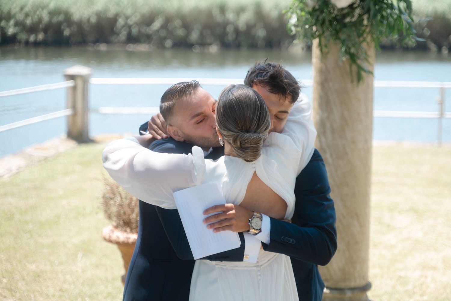 A group embraces in a heartfelt hug during an outdoor wedding ceremony near a lake.
