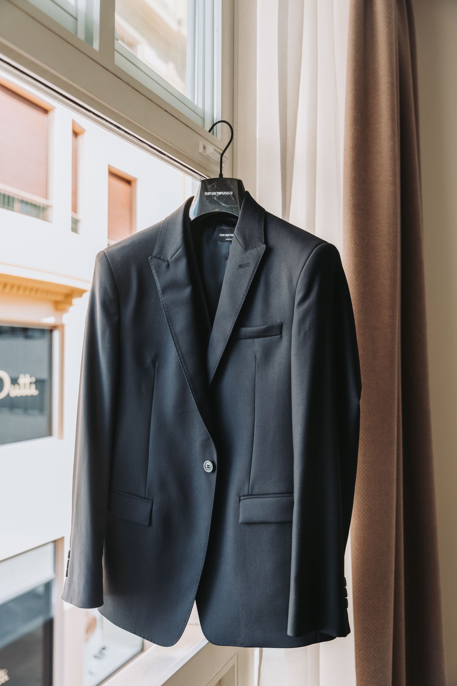 Black formal suit jackets hanging on wooden hangers against a beige curtained wall.