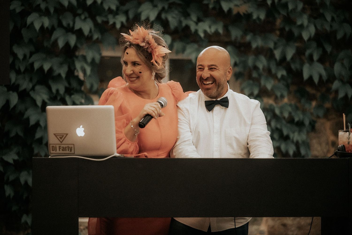 A couple sits together outdoors with a laptop and microphone against a leafy green backdrop at an event.