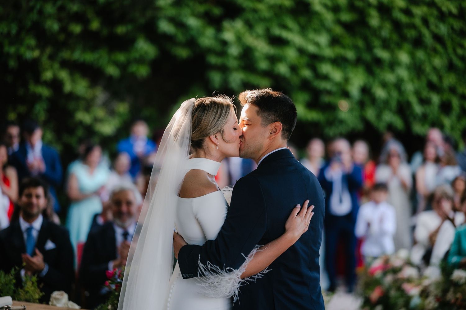 A couple shares an intimate moment during their wedding ceremony surrounded by greenery and guests.