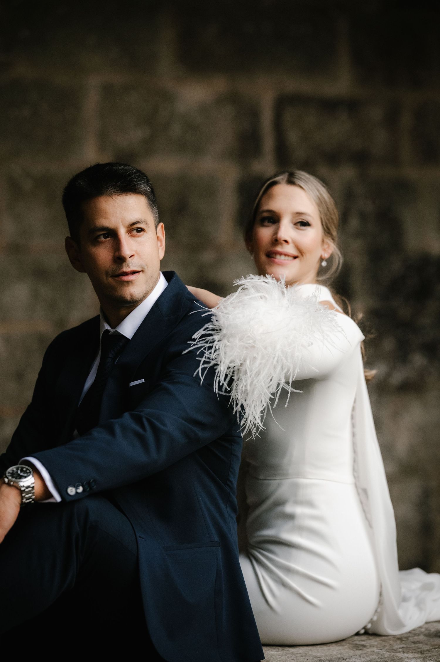 Elegant wedding couple poses against stone wall in classic white dress and navy suit.