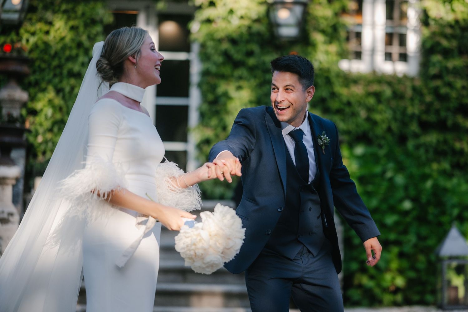 A joyful couple shares a candid moment during their outdoor wedding celebration with ivy-covered walls in the background.