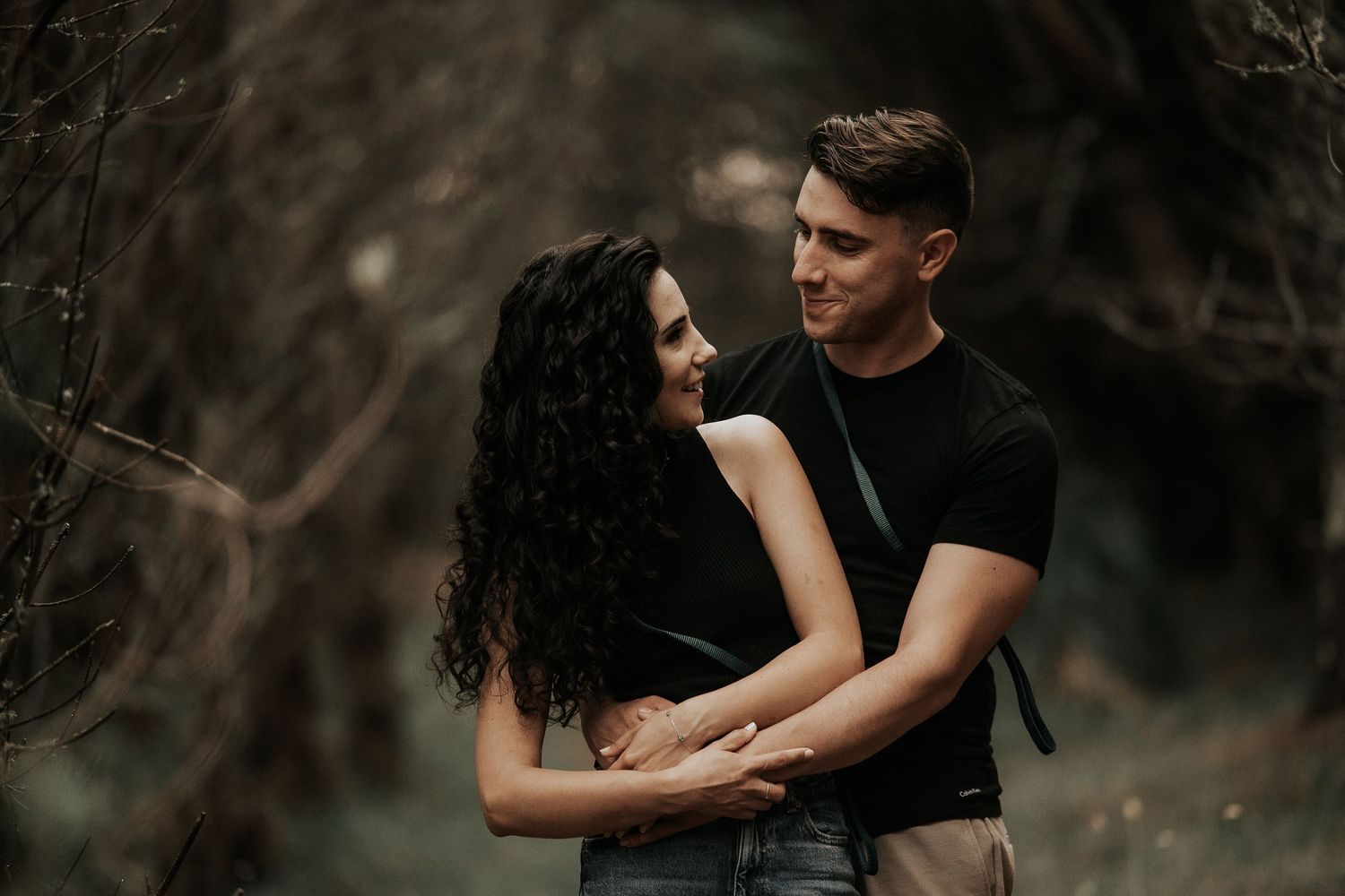 A romantic couple embraces in a moody forest setting with dark tones and atmospheric lighting.